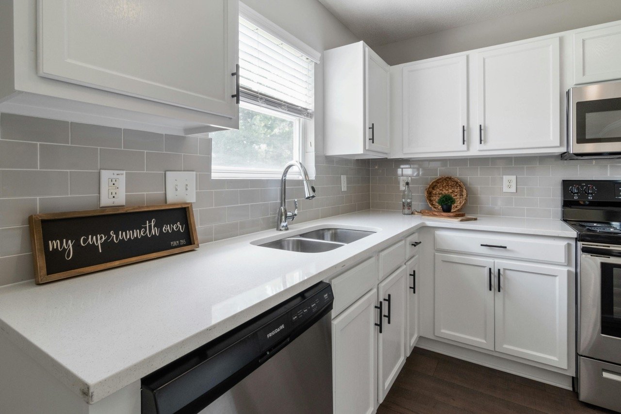 Kitchen with white cabinets, gray tile backsplash, window above sink, black dishwasher, stainless steel oven and microwave, decorative basket and plant on countertop.