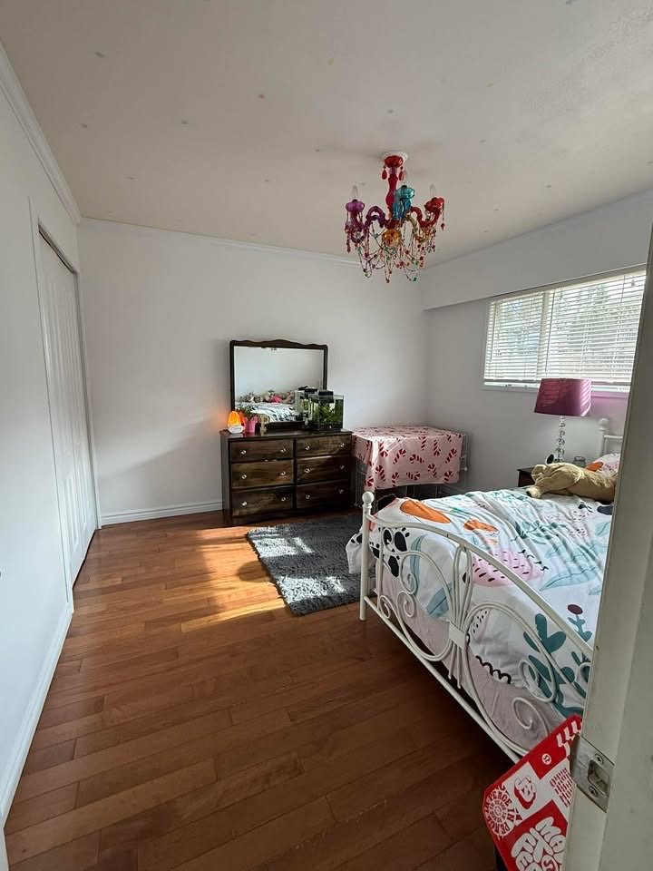 A bedroom with a white metal bed, colorful bedding, a pink lamp on a nightstand, a dresser with a mirror, a fish tank, and a window with blinds. There is a chandelier hanging from the ceiling and a small rug on the wood floor.
