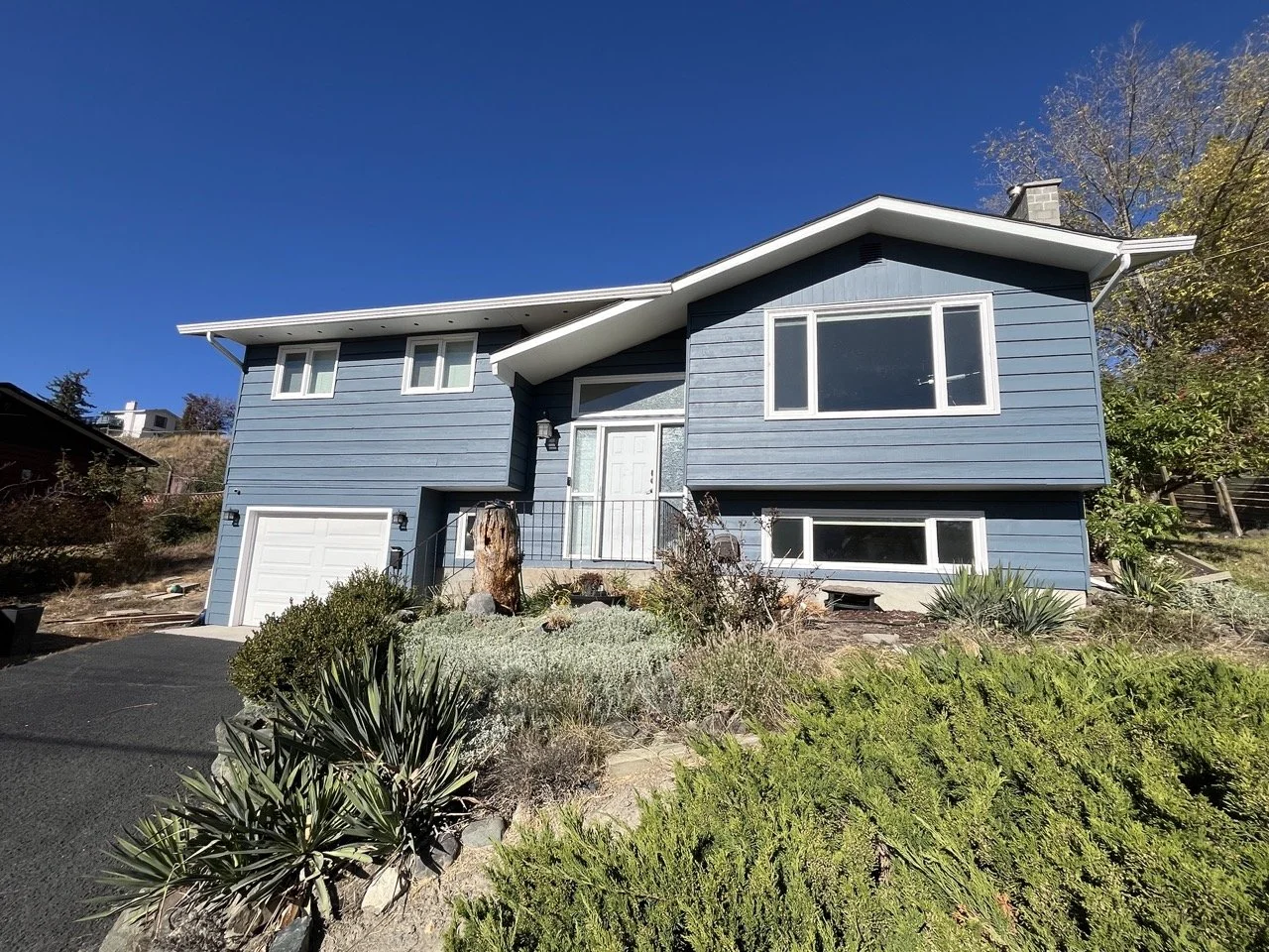 A blue, two-story house with white trim and multiple windows, situated on a sloped landscape under a clear blue sky, with greenery and shrubs in the front yard.
