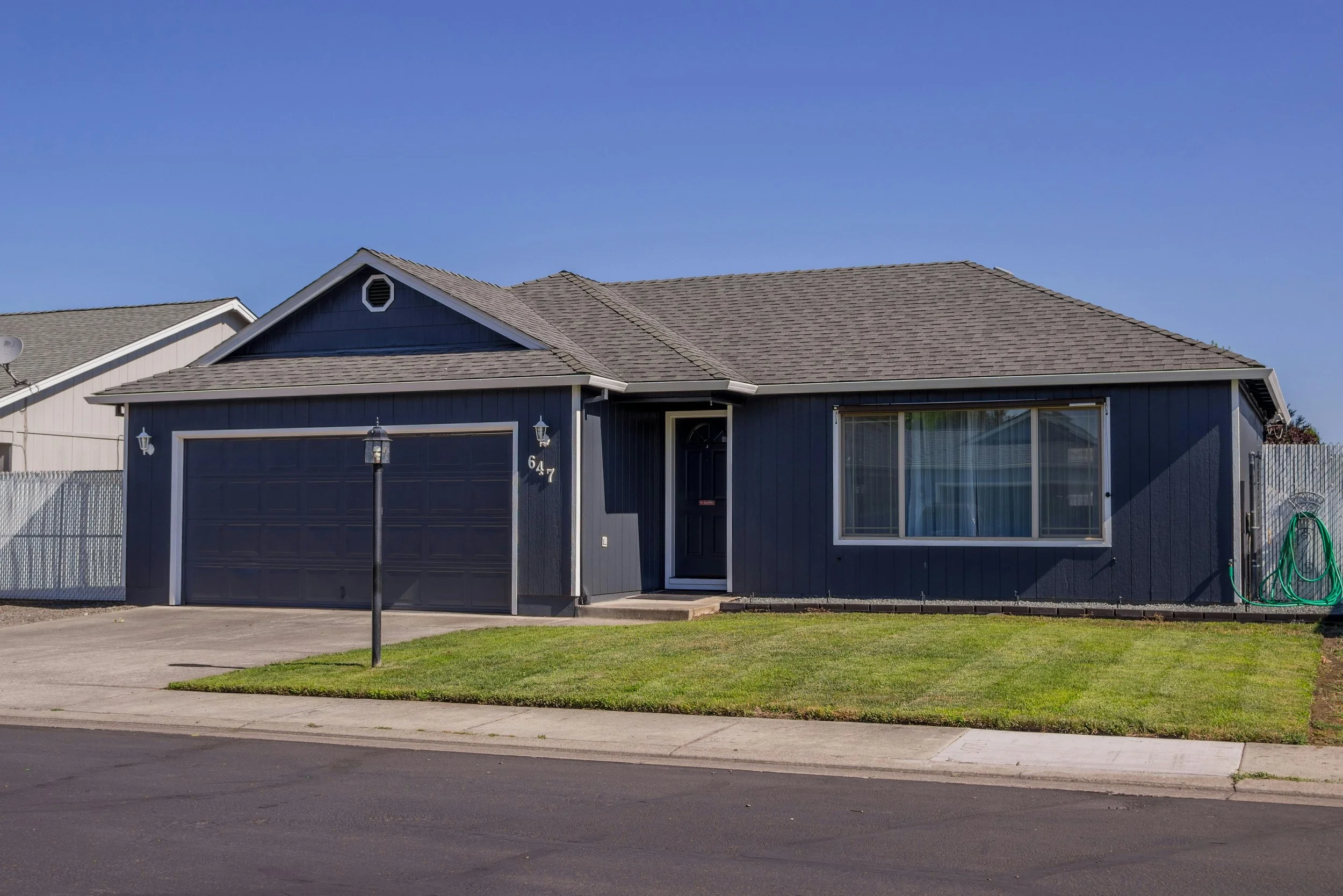 A single-story house with dark blue siding, a gray shingle roof, and a detached garage. There is a small front lawn, a paved walkway, a street lamp, and a hose reel on the side of the house. The house has a front door and large window with curtains.