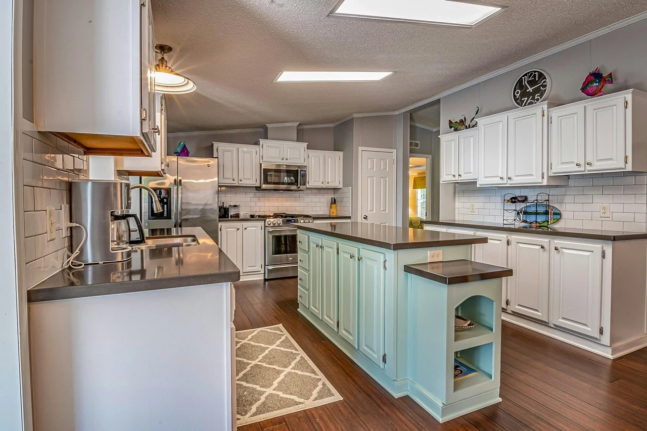 Kitchen with white cabinets, a stainless steel refrigerator, microwave, and stove, dark countertops, a beige and white geometric rug, and decorations on top of upper cabinets including a colorful fish sculpture, a clock, and a wooden bird.