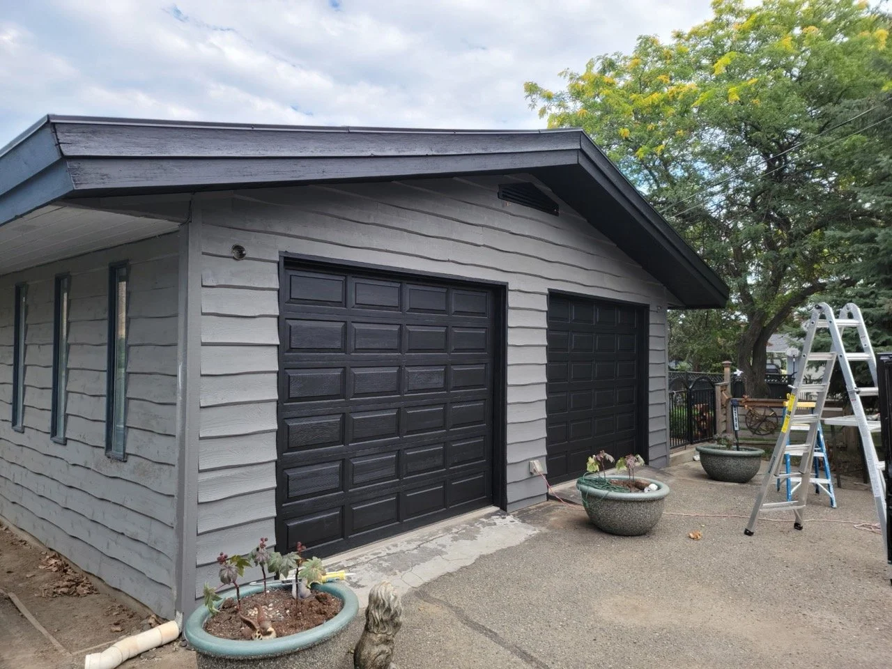 A two-car garage with gray siding, black garage doors, and a sloped roof. There are two large planters with plants in front and a ladder on the right side. A tree with green leaves is in the background.