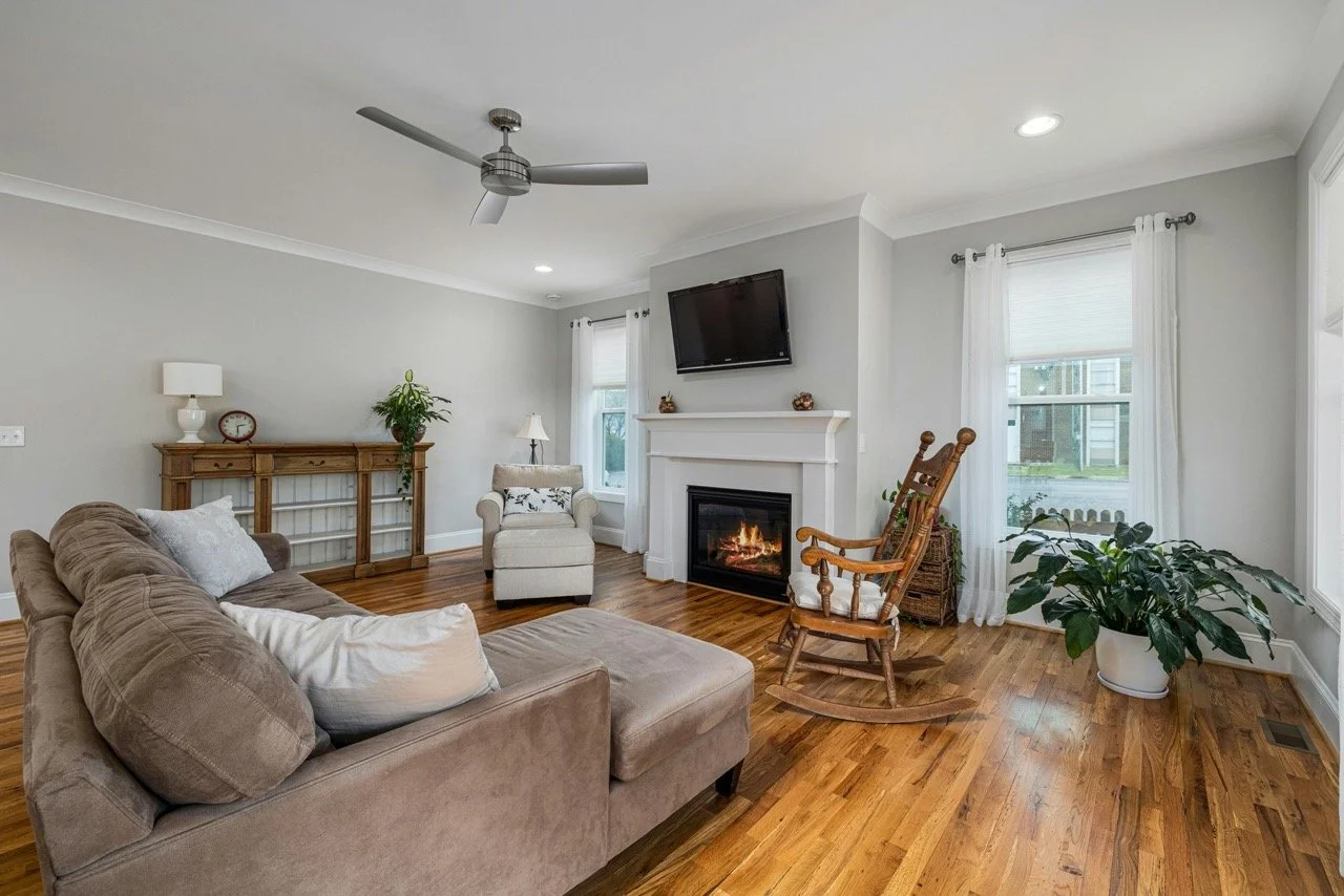Living room with beige sofa, wooden rocking chair, beige armchair, fireplace, wall-mounted TV, hardwood floor, large window with white curtains, potted plants, ceiling fan, and recessed ceiling light.