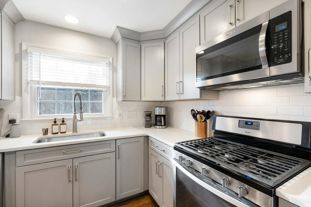 Kitchen with white cabinets, stainless steel microwave, stove, coffee maker, utensil holder, window with blinds, sink, and countertop