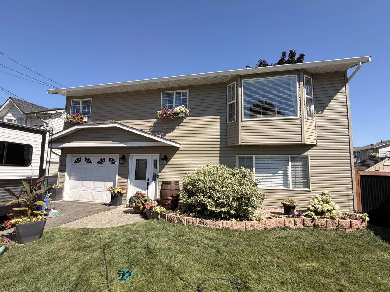 Front view of a two-story beige house with a garage, bay window, and flower beds with colorful flowers in pots and around the garden, under a clear blue sky.