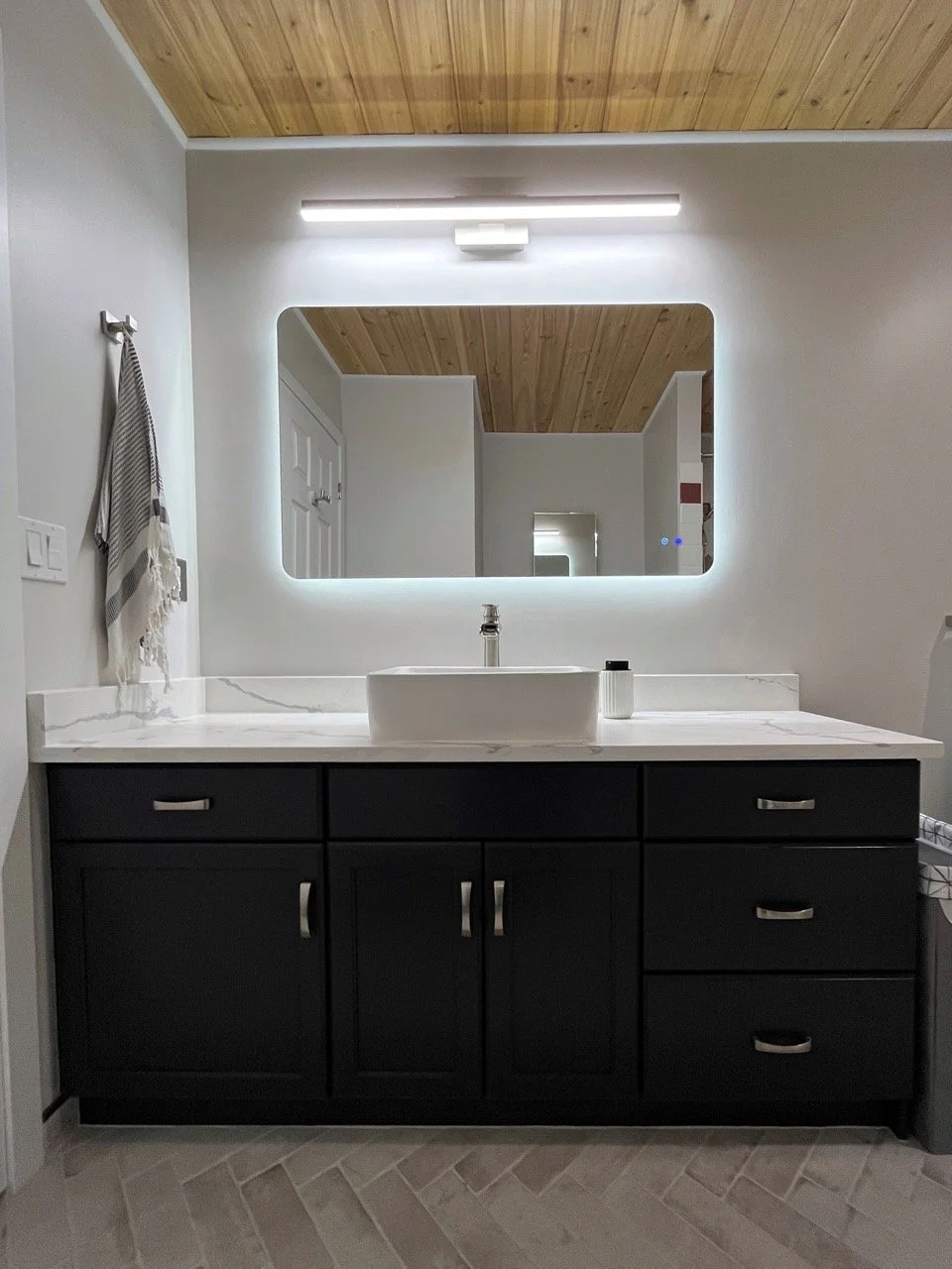 Modern bathroom vanity with a black cabinet, white marble countertop, rectangular vessel sink, large backlit mirror, wooden ceiling, towel hanging on the wall, and a light fixture above the mirror.