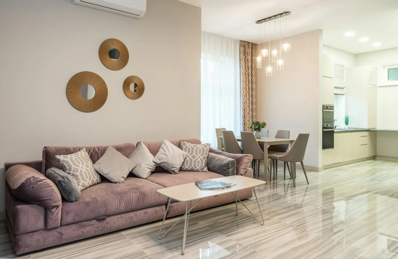 Living room with pink sofa, decorative pillows, wall mirrors, coffee table, dining table with chairs, and a kitchen area in the background with white cabinets and countertops.