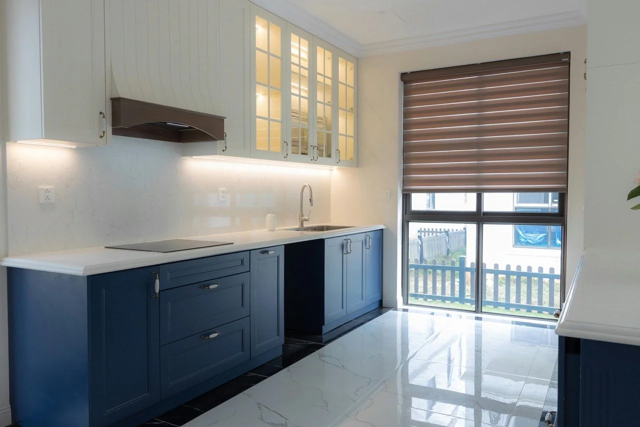 Modern kitchen with blue lower cabinets, cream upper cabinets, a white countertop, and a large window with brown blind outside. Includes a stovetop and a sink.