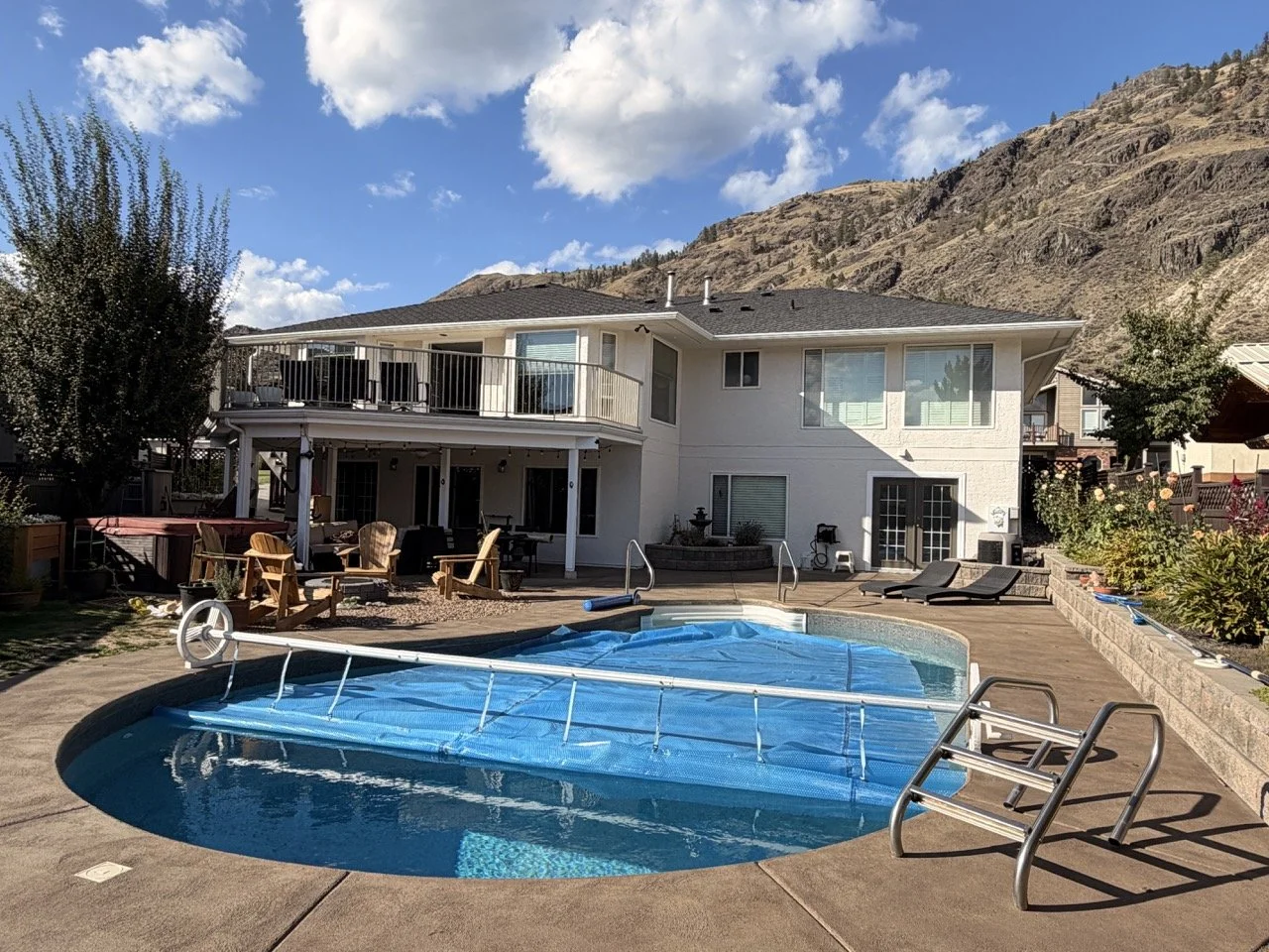 Backyard with swimming pool covered by a blue pool cover, lounge chairs, patio furniture, and a two-story house with a balcony, surrounding mountains, and blue skies.