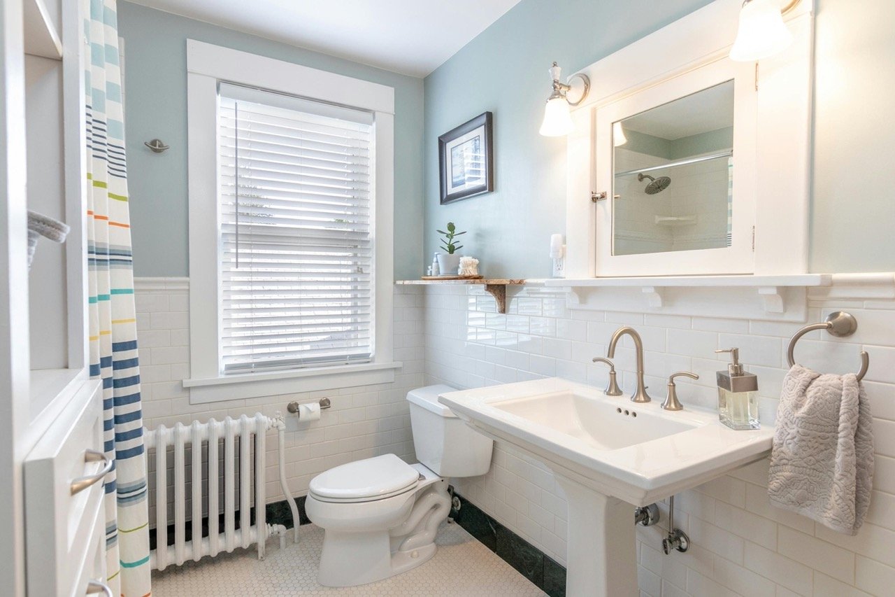 Bright bathroom with light blue walls, white subway tile accents, a window with blinds, a white toilet, a pedestal sink, a mirror with light fixture, a small wall shelf with decorative items, a framed picture, and a striped shower curtain.