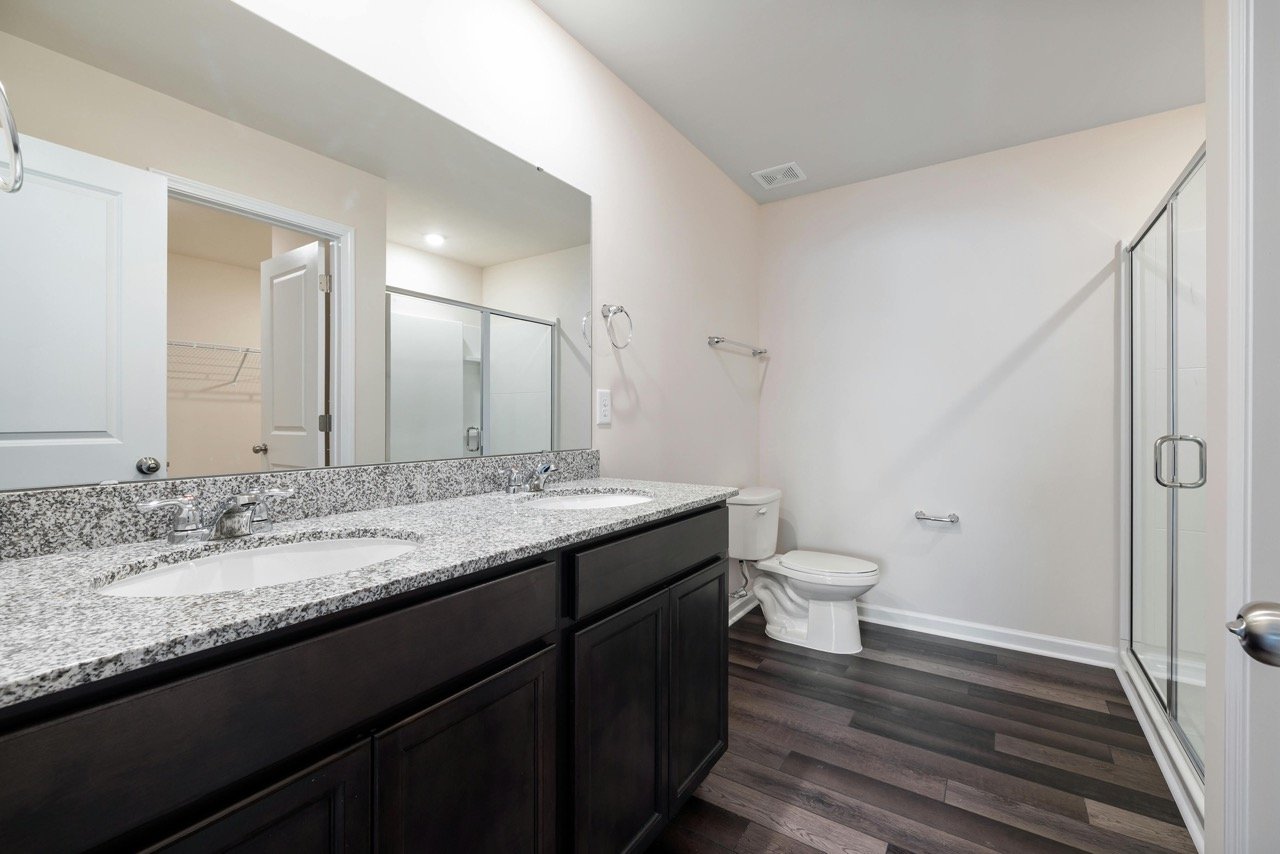 Bathroom with double sink vanity, granite countertop, large mirror, toilet, glass shower enclosure, and wood-look flooring.