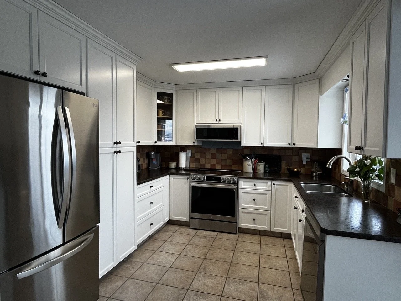 Kitchen with white cabinets, black countertops, stainless steel refrigerator, stove, microwave, and a window with a plant.