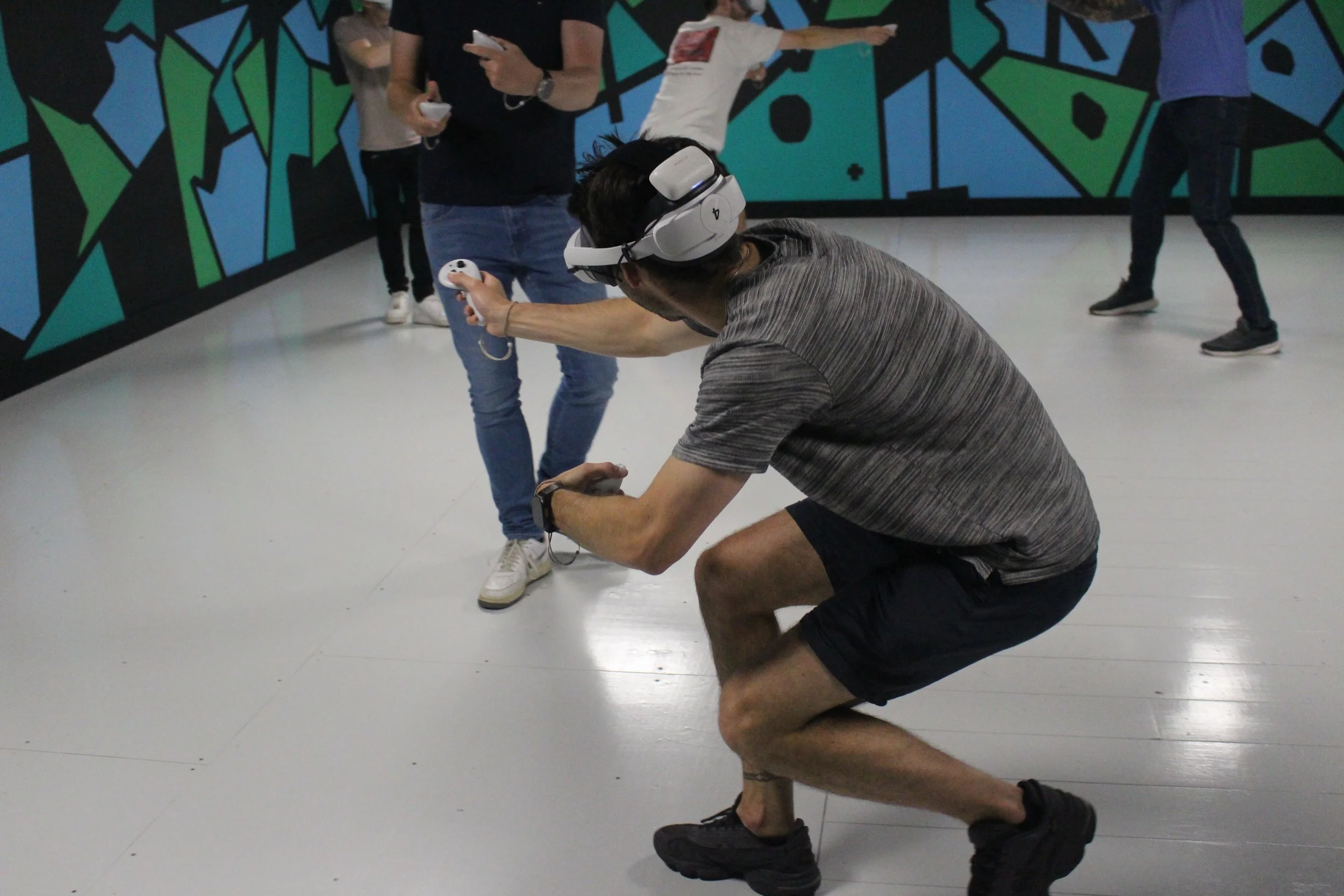 A man kneeling and wearing a virtual reality headset and holding a VR controller, engaging in a virtual experience with other people in the background at an indoor venue with a colorful geometric wall design.