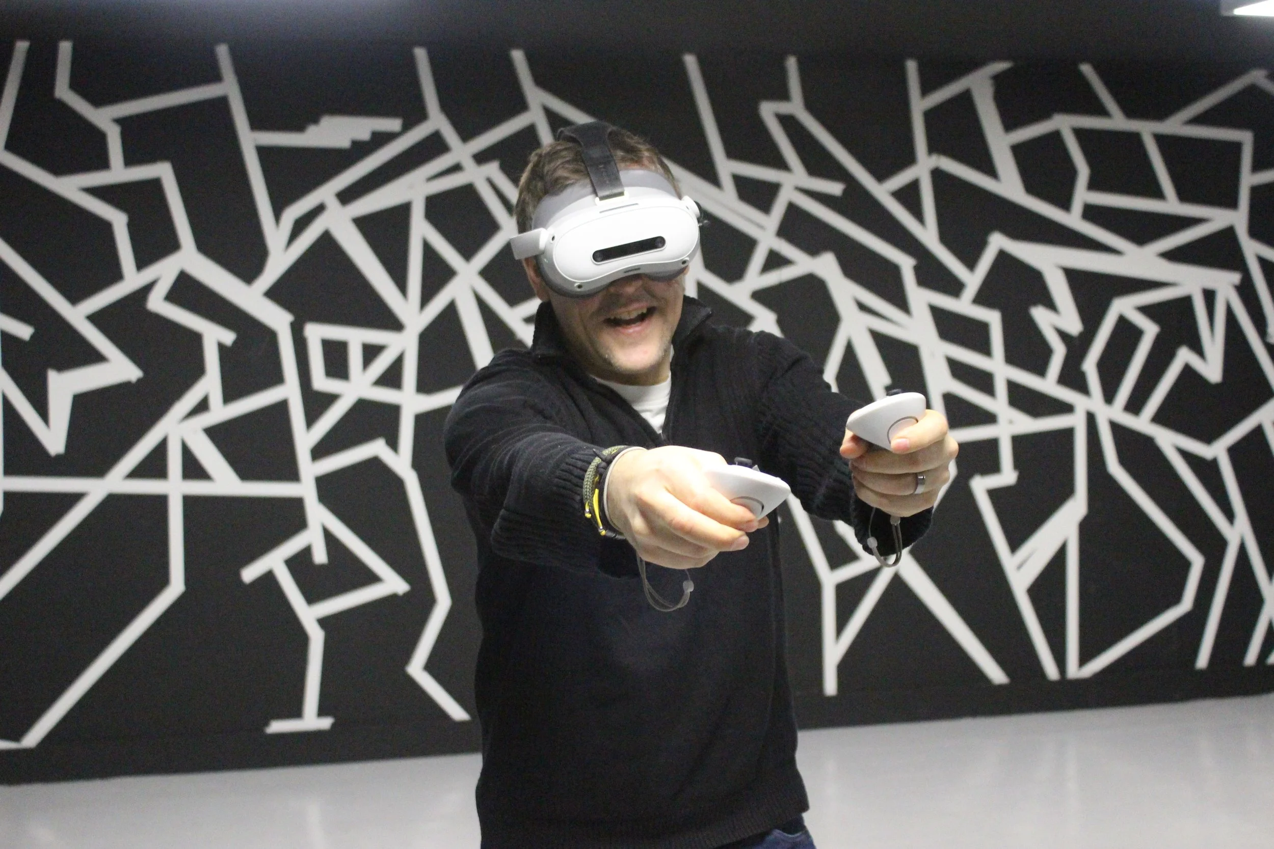 A man wearing a virtual reality headset and holding a controller in each hand, smiling and experiencing virtual reality in a room with black and white geometric wall art.