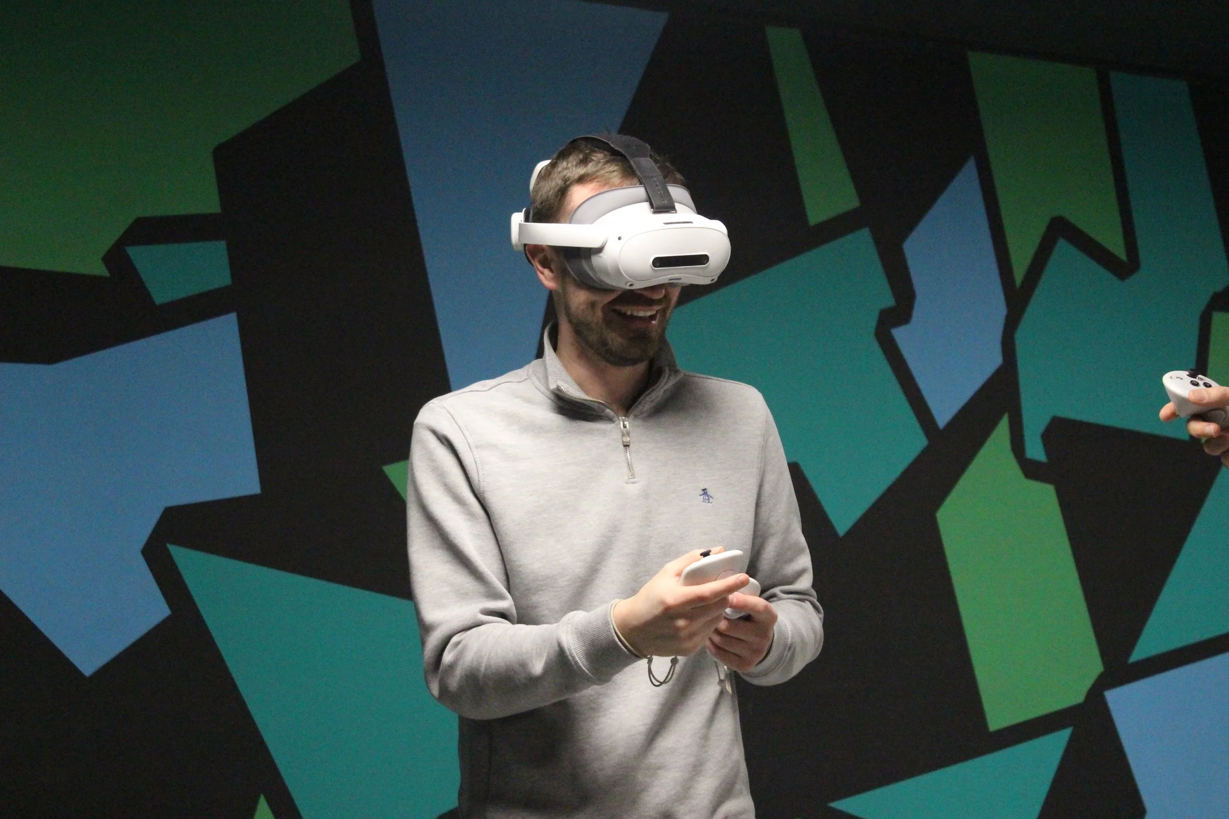 A man wearing a virtual reality headset and holding a game controller, standing in front of a colorful geometric mural.
