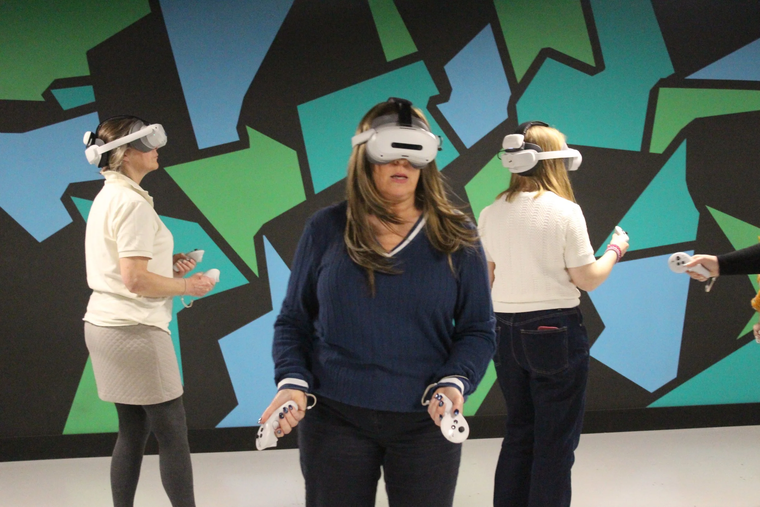 Four women wearing virtual reality headsets and holding controllers in an indoor setting with a colorful, geometric mural on the wall.