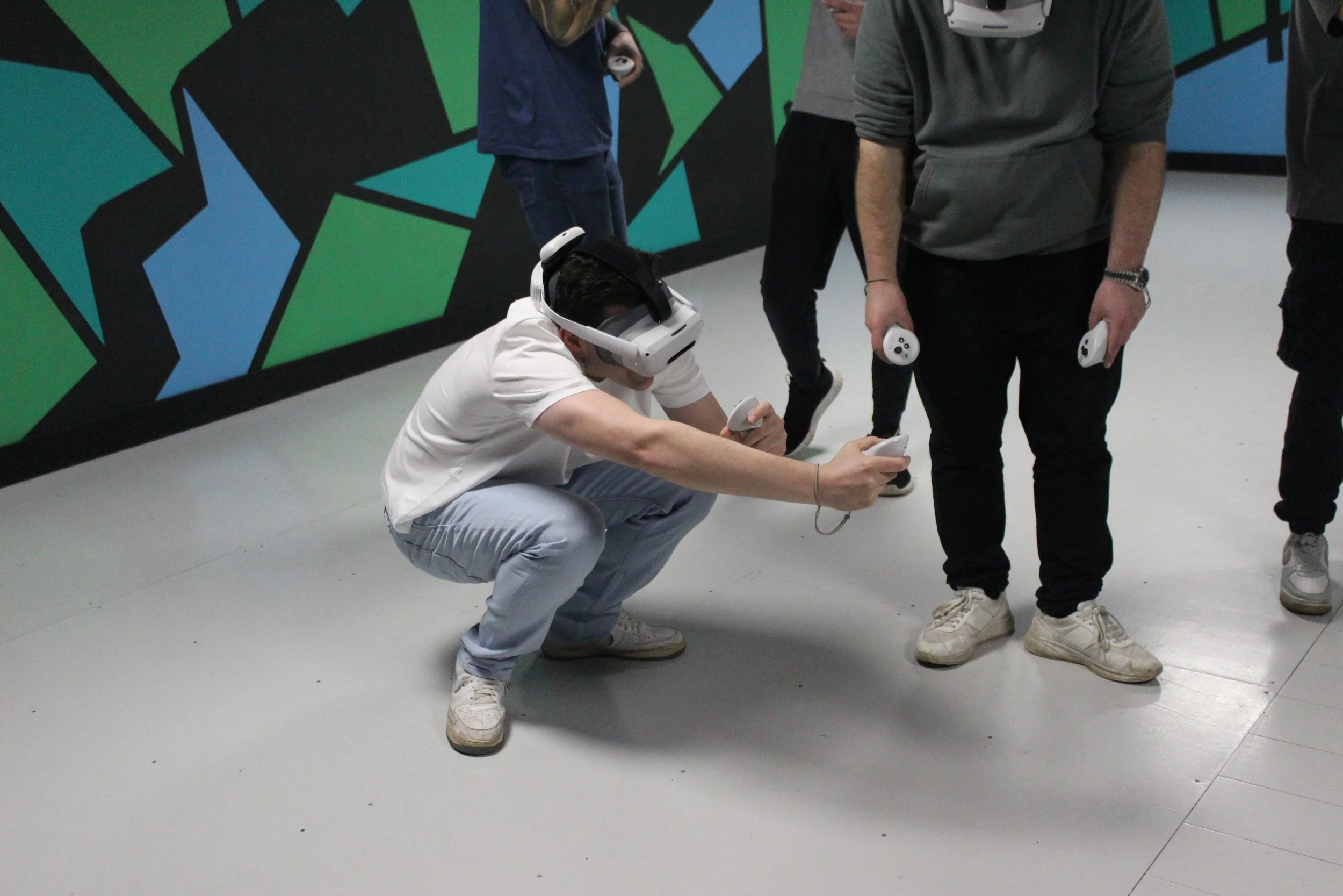 Person in white shirt and light gray pants crouching while holding a VR controller, wearing VR headset in a room with colorful geometric wall art, and other people holding VR controllers around him.
