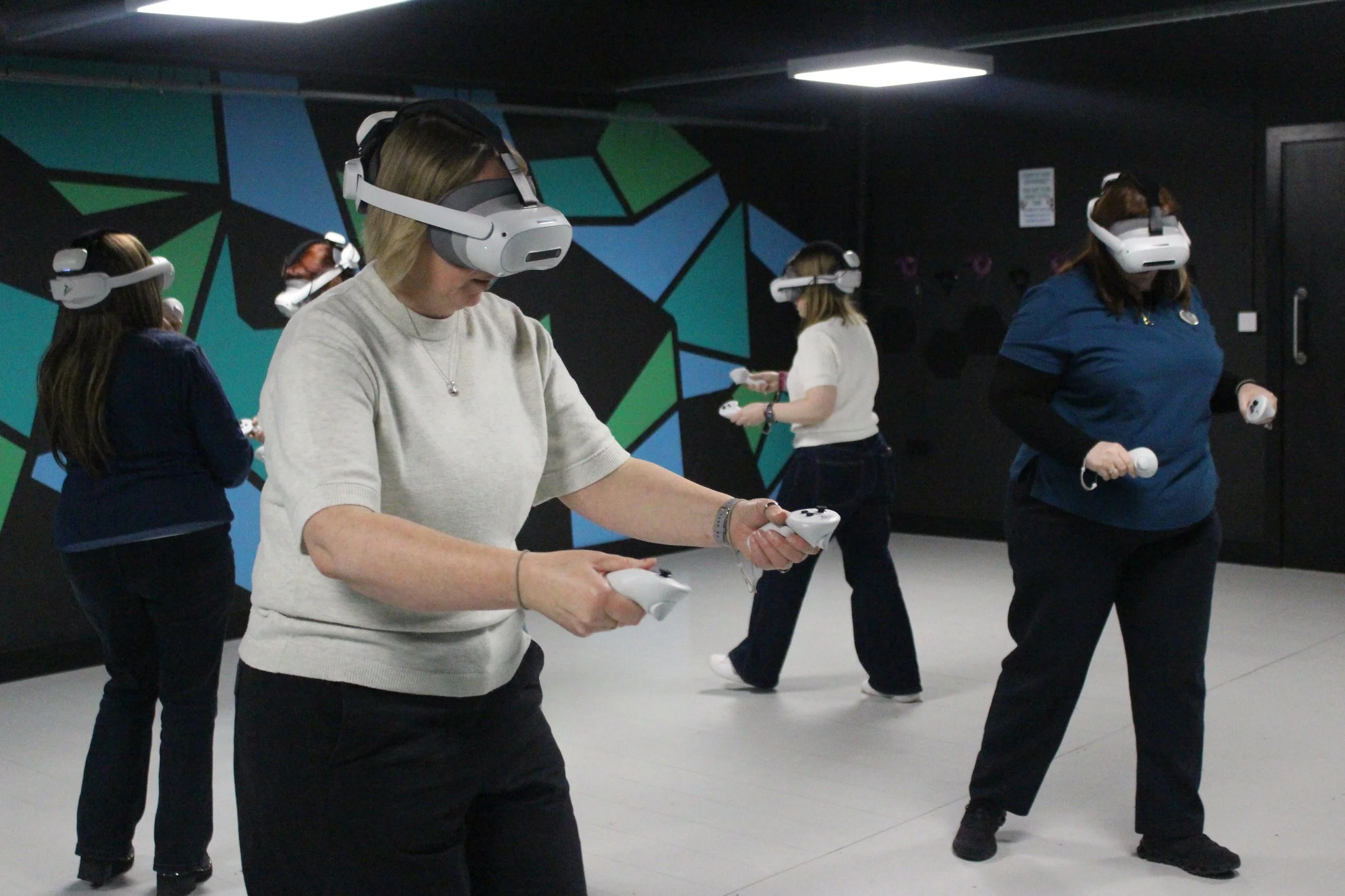 Five women wearing virtual reality headsets and holding controllers in a room with a black wall and colorful geometric mural.
