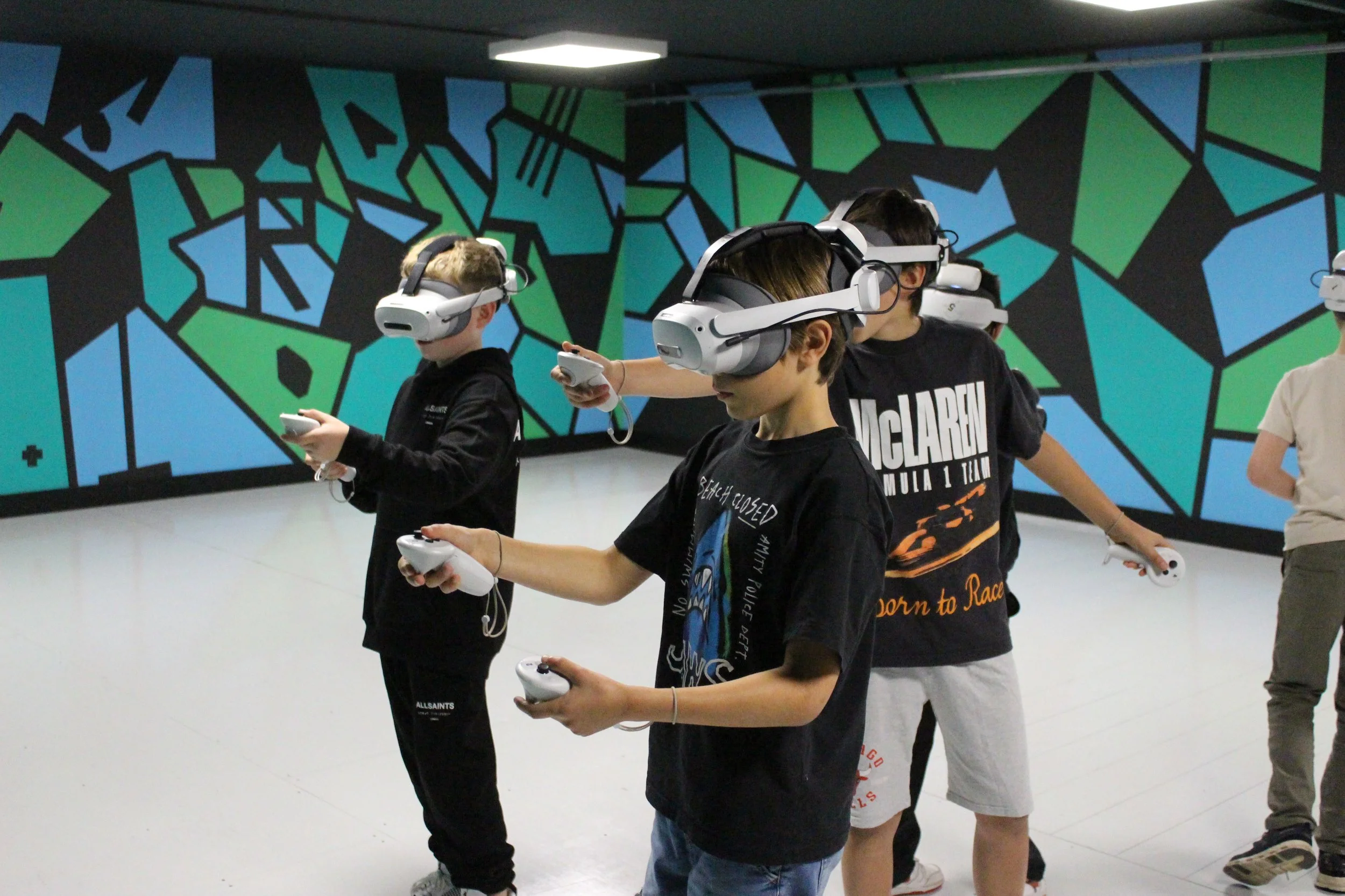 Young people wearing virtual reality headsets and holding controllers in a room with a colorful, geometrically patterned mural on the wall.