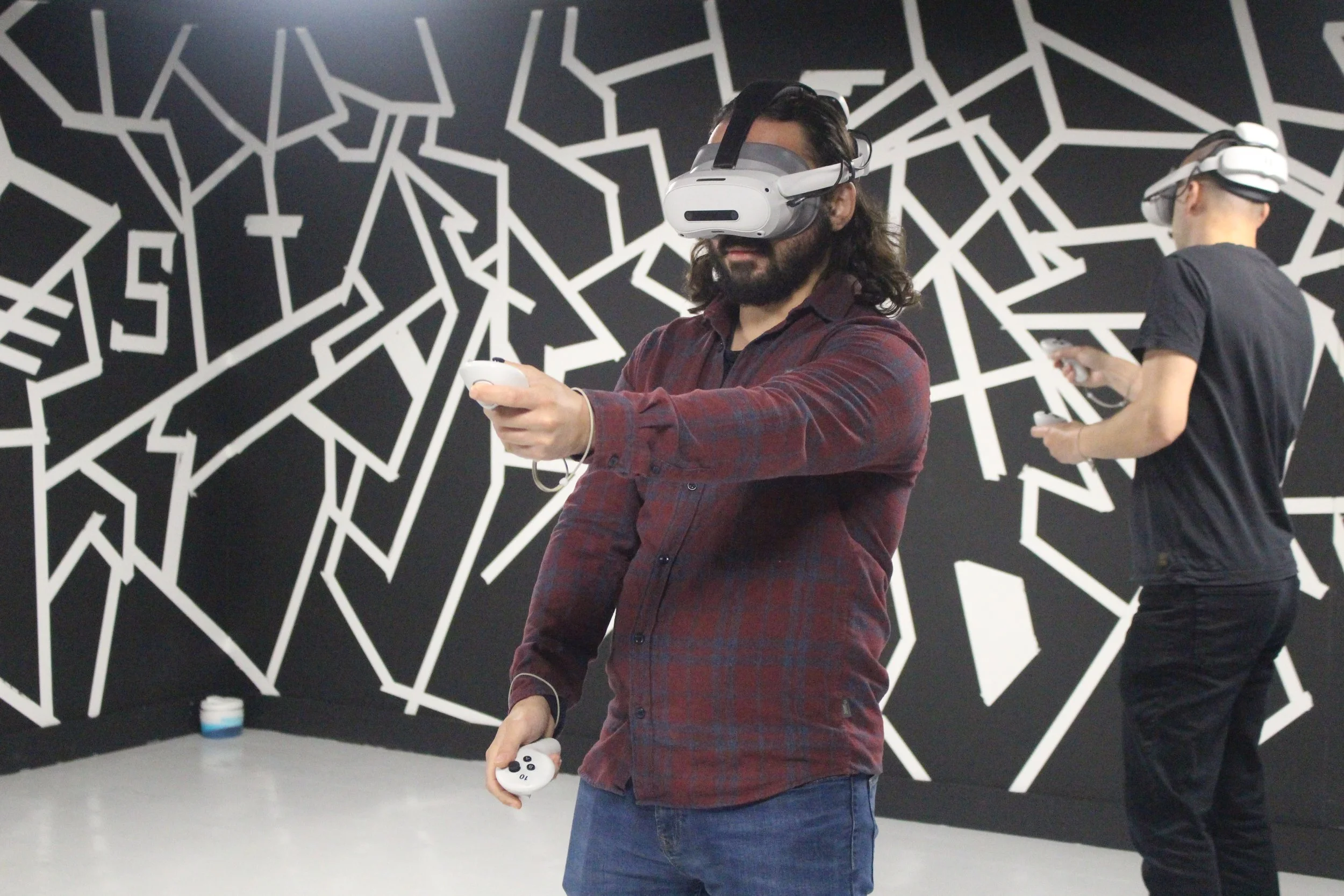 Two men using virtual reality headsets in a room with black and white geometric wall art, one holding a game controller and the other looking at his phone.