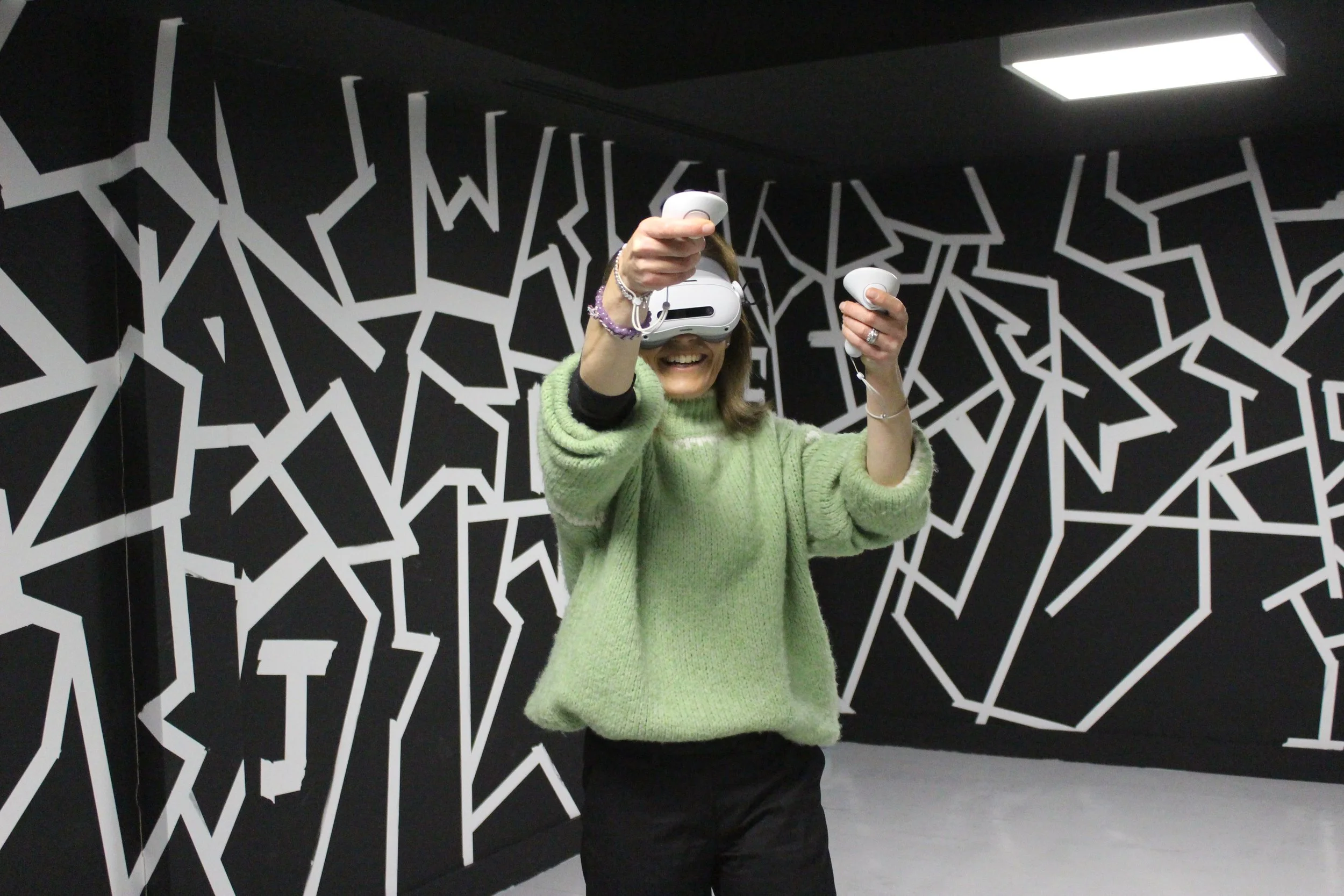 Woman wearing a virtual reality headset and holding controllers, smiling while standing in a room with black and white abstract wall art.