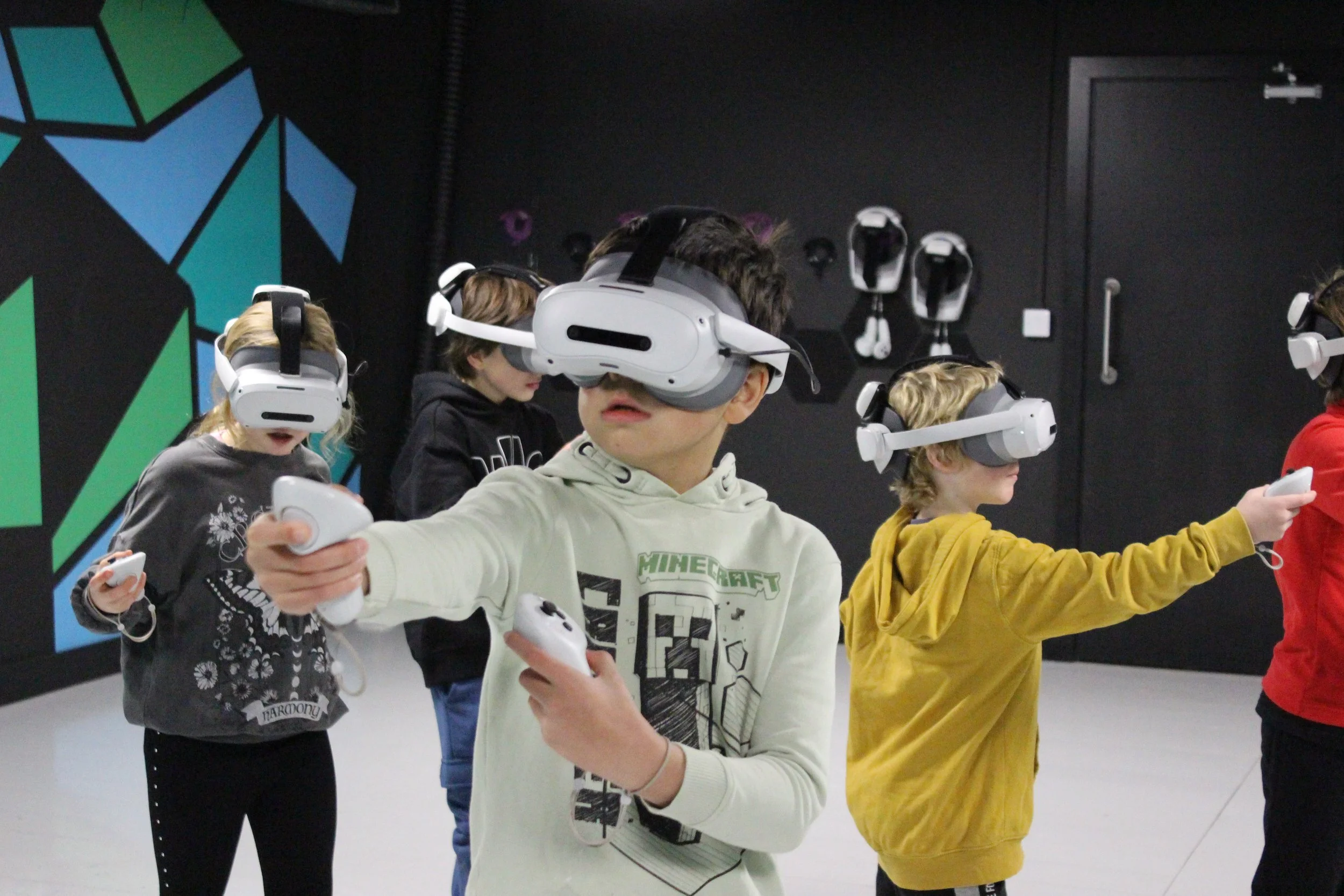 Children wearing VR headsets and holding controllers in a black indoor space with colorful geometric wall art.