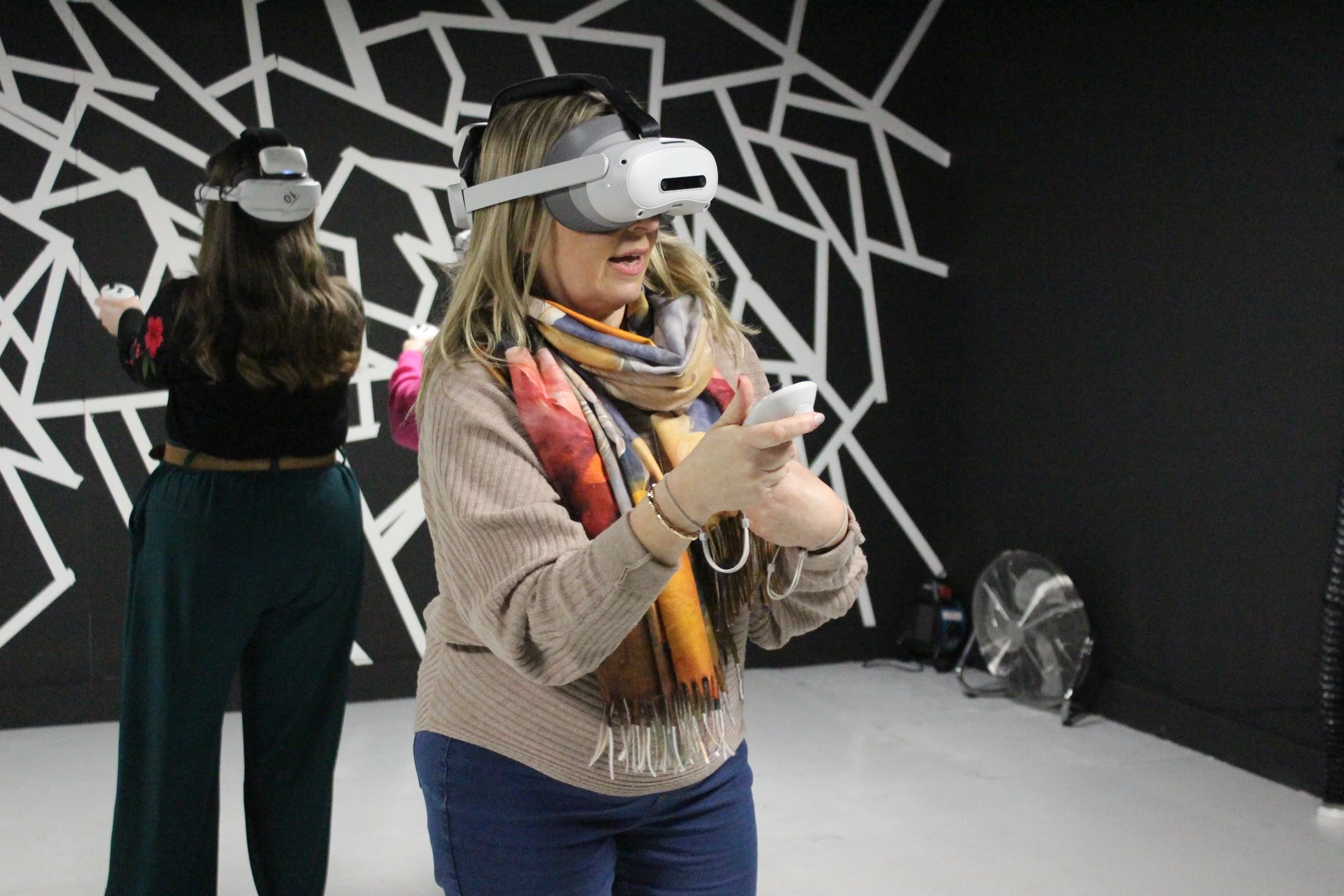 Women wearing virtual reality headsets in an indoor space with a black and white geometric mural on the wall.