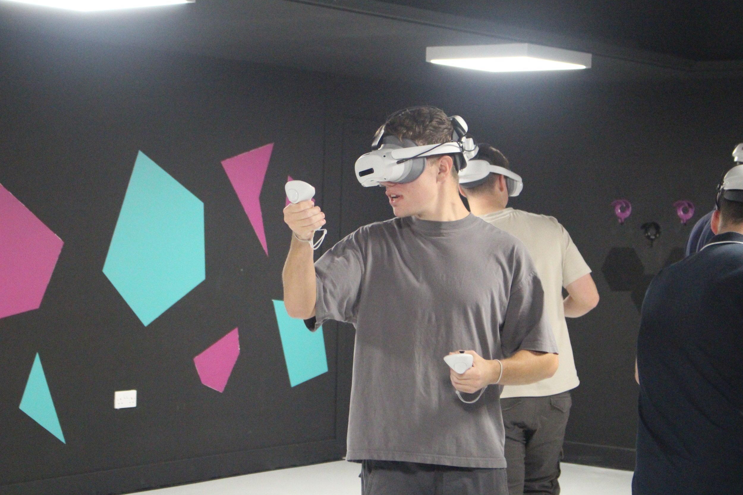 Young man wearing a virtual reality headset holding a VR controller in one hand and a small device in the other, with other people in the background also wearing VR headsets. The room has a black wall with colorful geometric shapes.