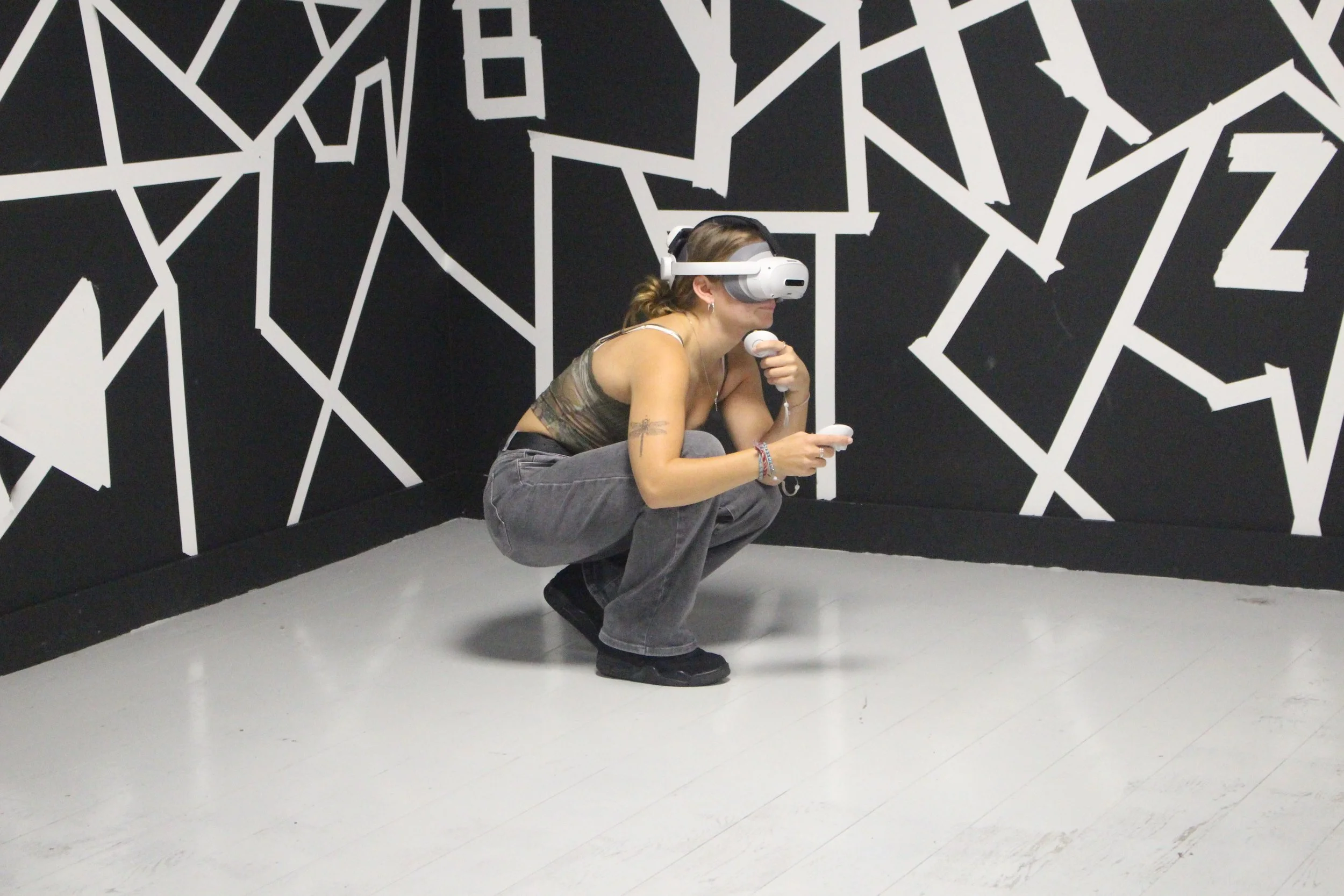 A woman crouching on the floor using a virtual reality headset and controllers in front of a black and white geometric wall.