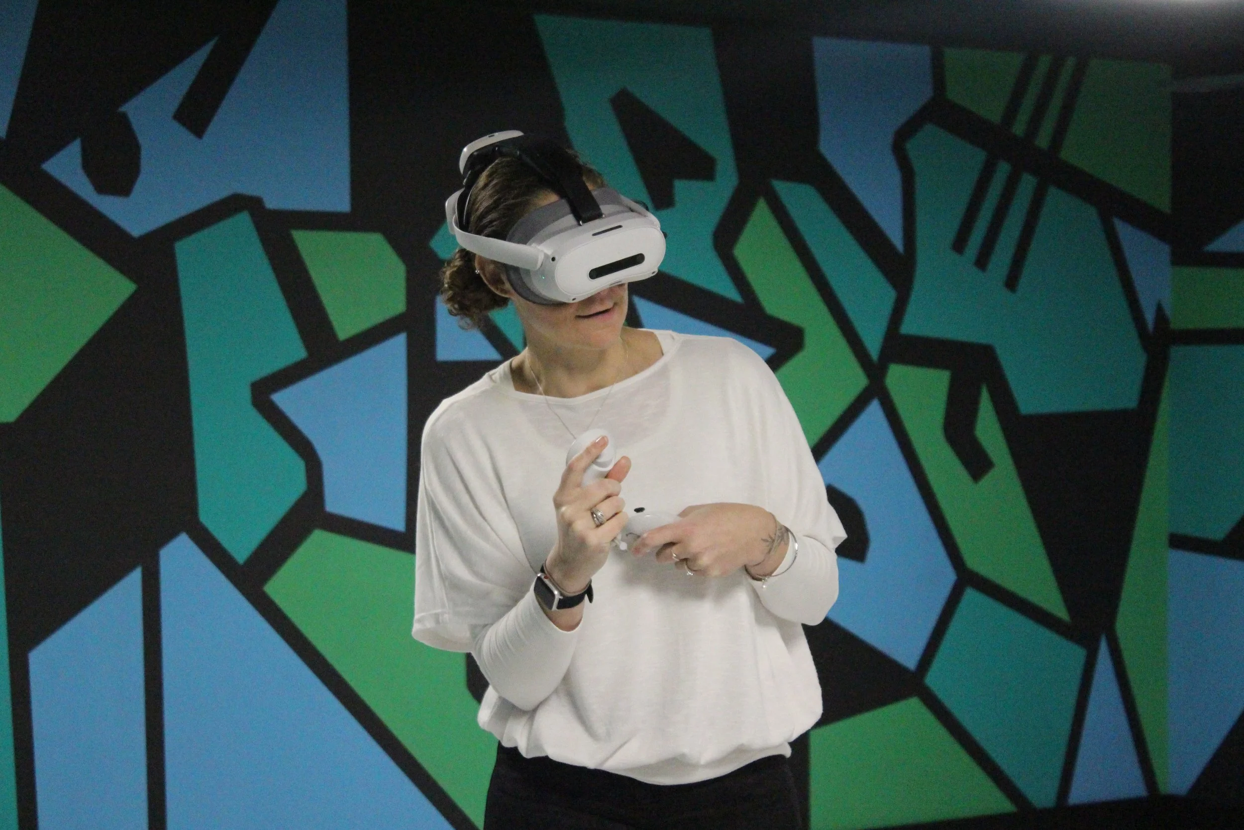 A woman wearing a virtual reality headset holding a VR controller against a colorful geometric mural background.