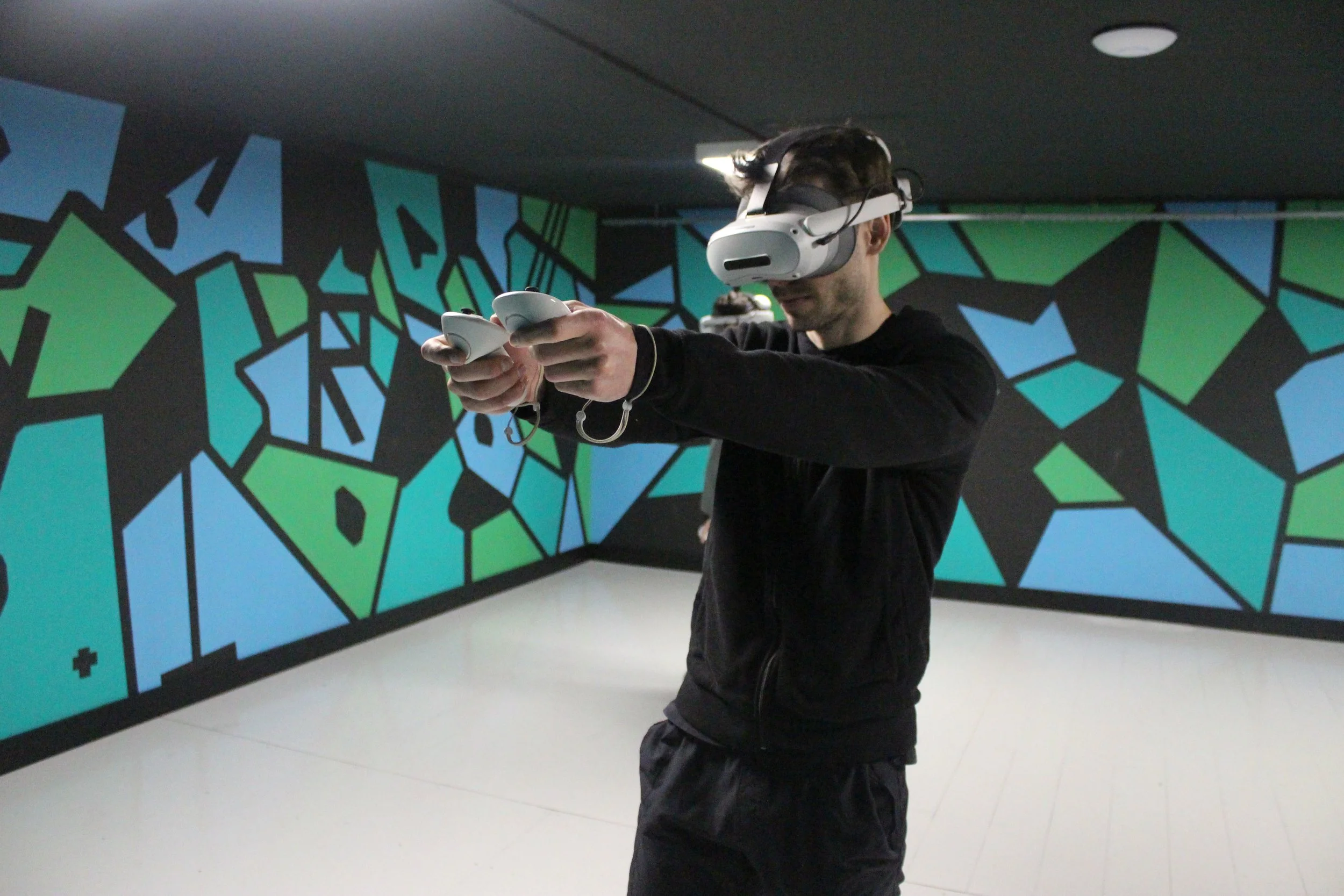 A man wearing a virtual reality headset and holding controllers in a room with geometric blue and green wall art.