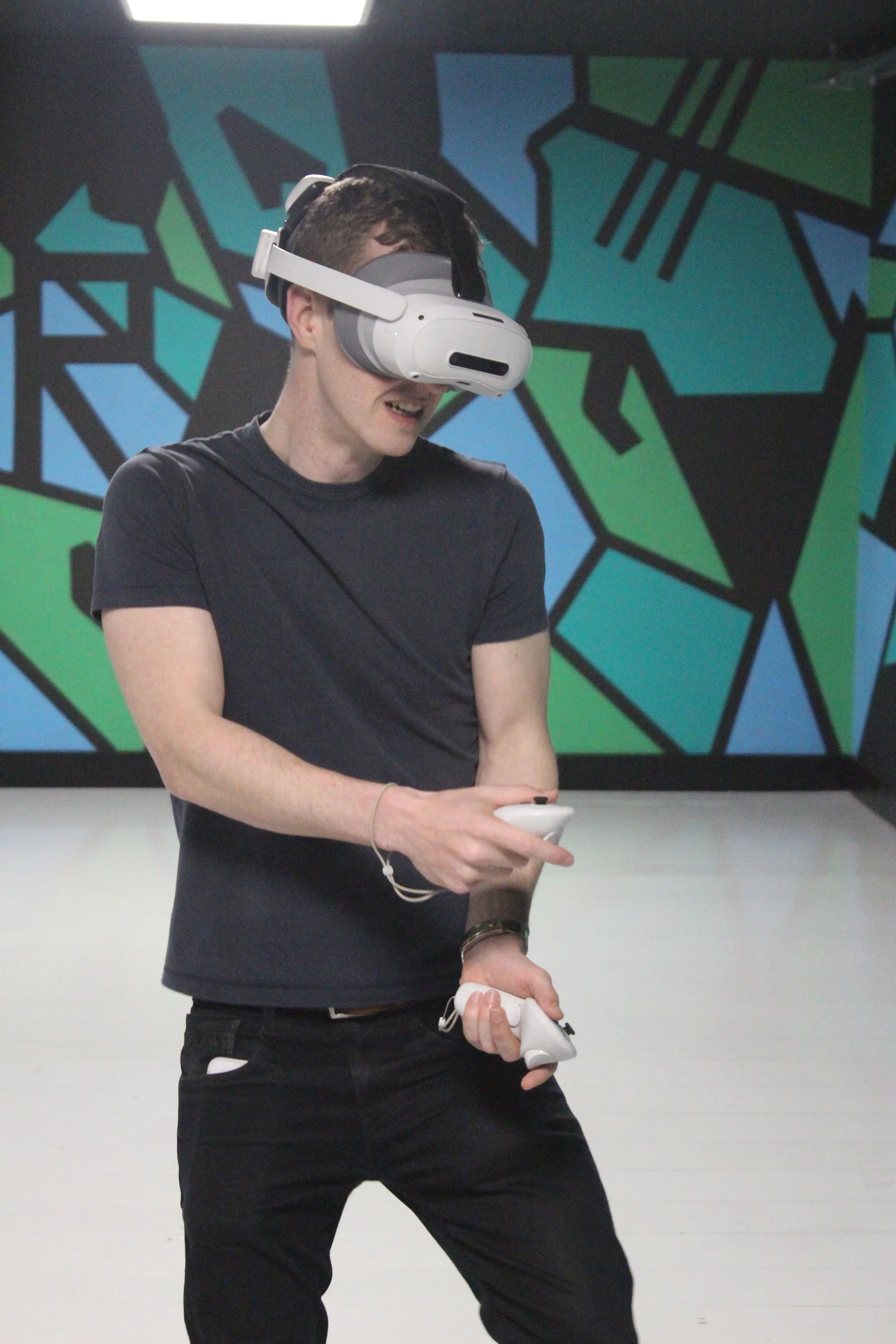 A young man in a black t-shirt and black pants wearing a virtual reality headset and holding VR controllers in an indoor space with colorful geometric wall art.