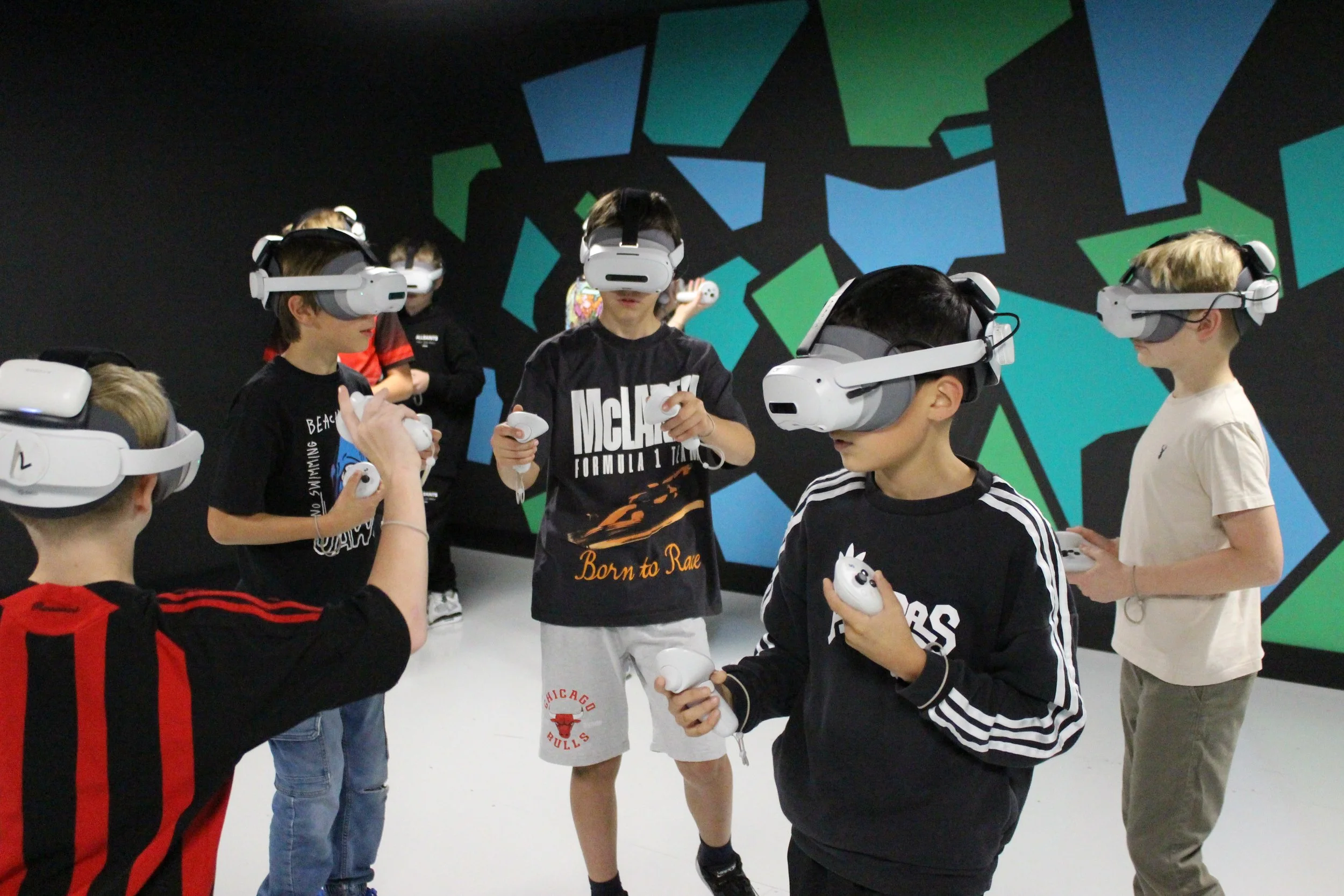 Group of children wearing virtual reality headsets and holding controllers in a room with colorful geometric wall art.