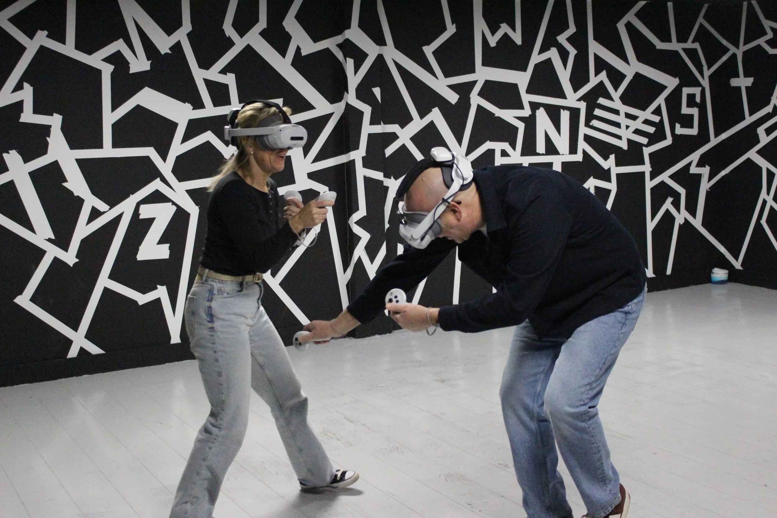 Two people wearing virtual reality headsets are interacting with VR controllers in a room with black and white geometric wall art.