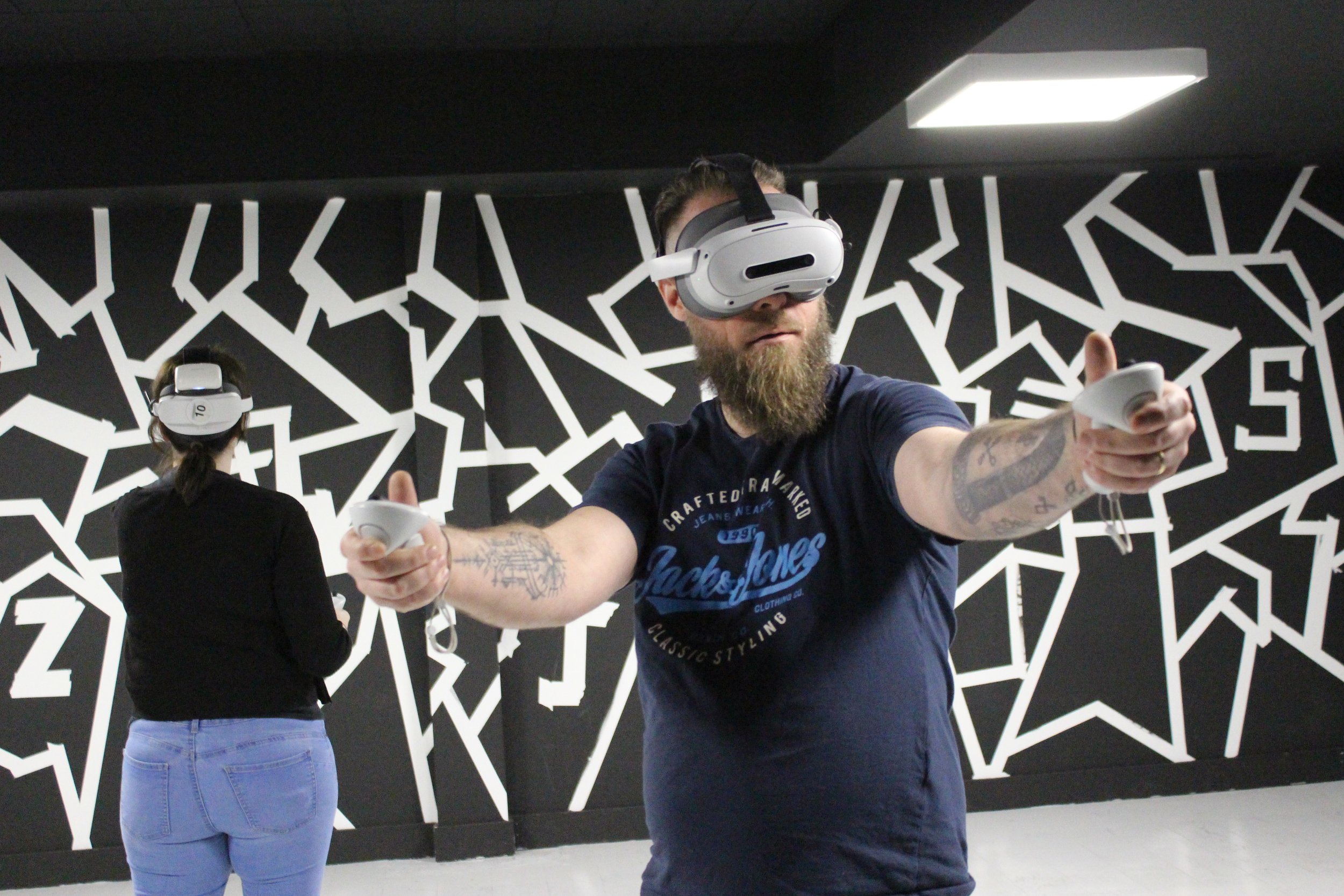 A man with a beard and tattoos wearing a dark T-shirt using a virtual reality headset and hand controllers in a room with a black and white geometric patterned wall.