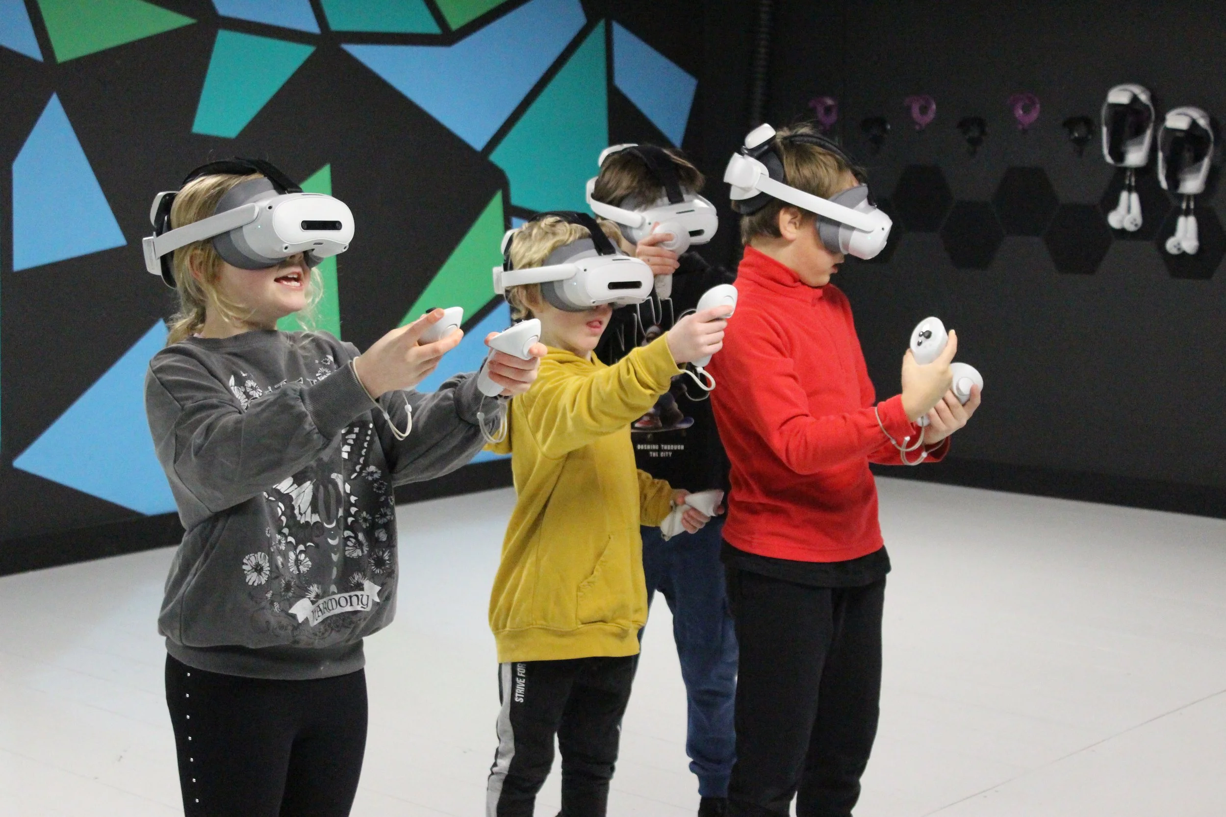 Four children wearing virtual reality headsets and holding controllers, standing in a room with a black wall decorated with colorful geometric shapes and additional VR headsets hanging on the wall.