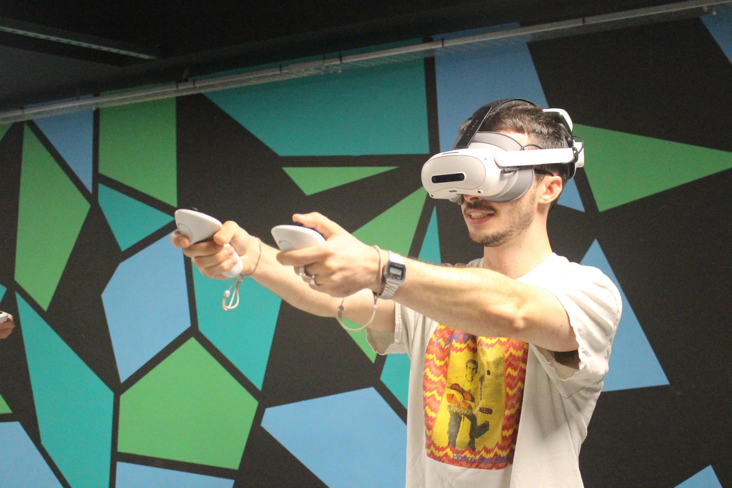 A man wearing virtual reality goggles and holding two game controllers, smiling while playing a VR game. The background features a modern, geometric mural in blue, green, and black.