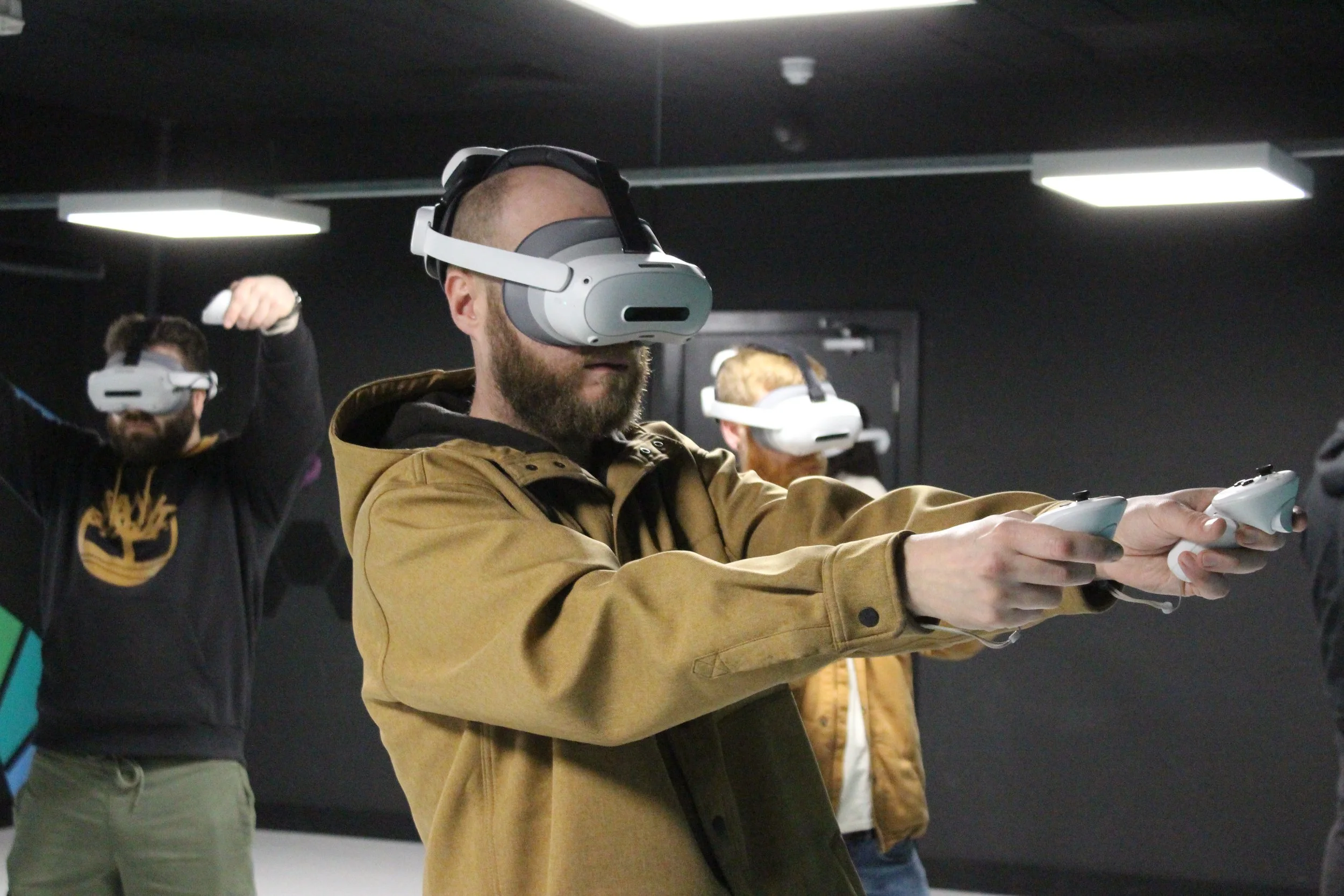 Three men in a dark room playing a virtual reality game with VR headsets and controllers, one in the foreground wearing a tan jacket.