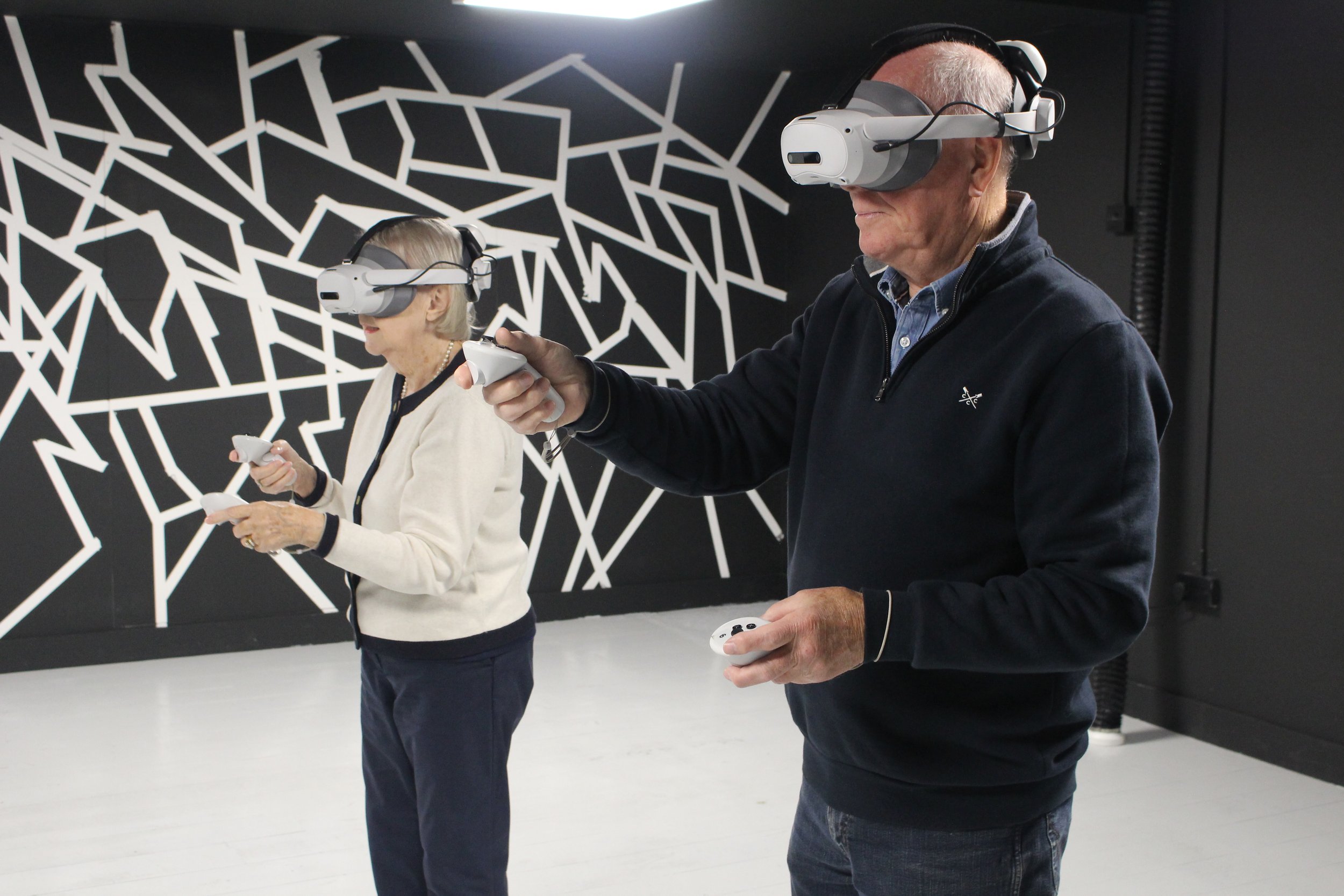 Two elderly individuals, a man and a woman, wearing virtual reality headsets and holding controllers in a room with black and white geometric wall art.