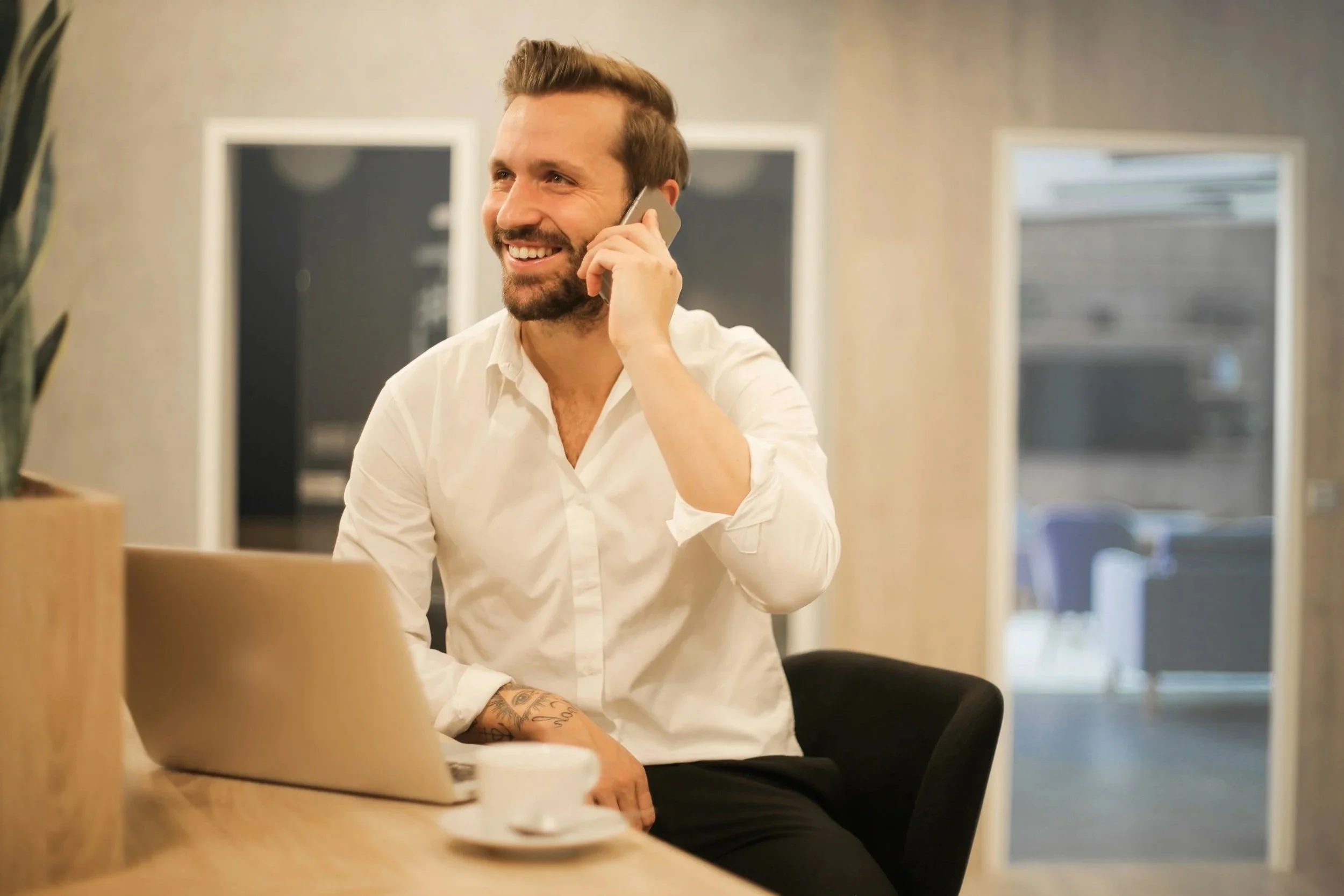 A smiling man with a beard and tattoos on his arm, wearing a white shirt, sitting at a table with a laptop, talking on the phone in an office environment.