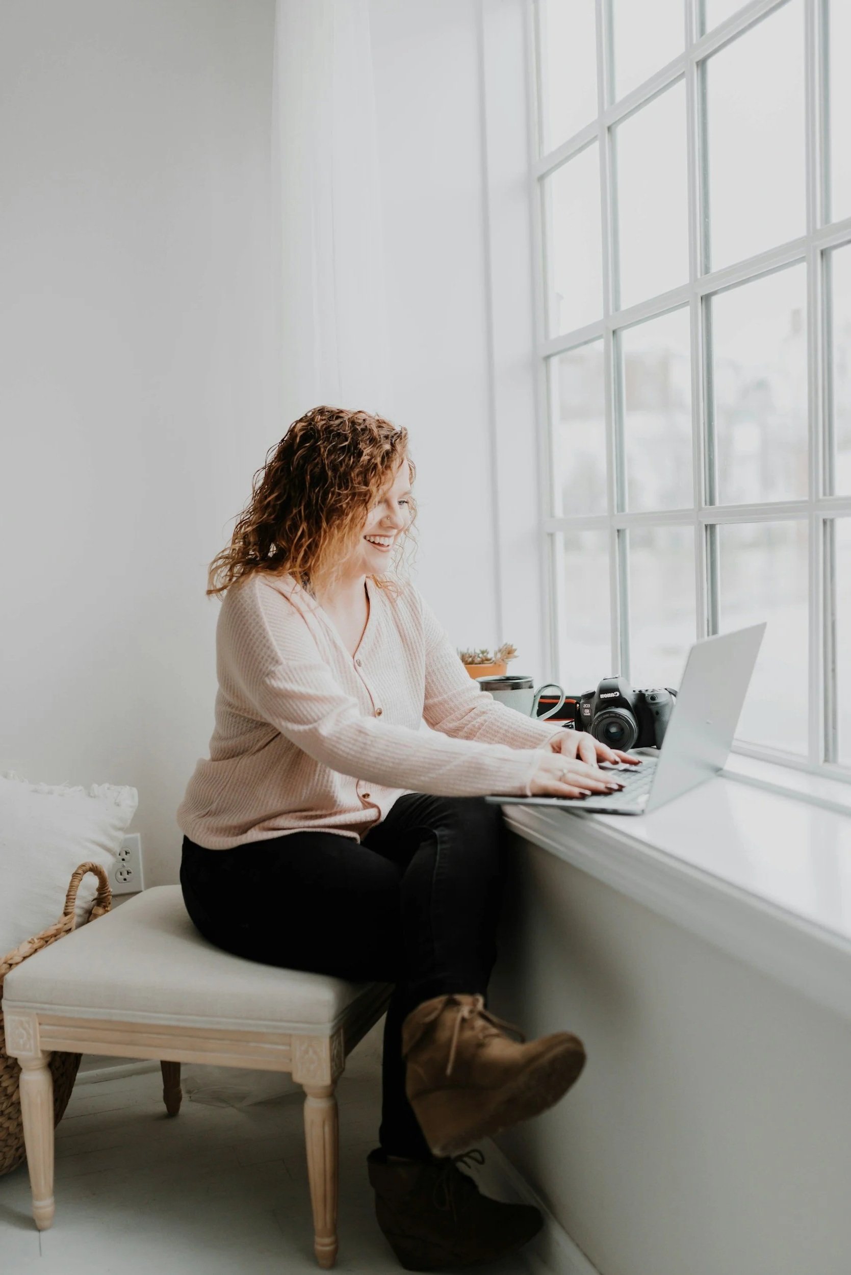 A woman sitting on a white cushioned chair at a window, using a laptop, with a camera and a cup on the windowsill, smiling.