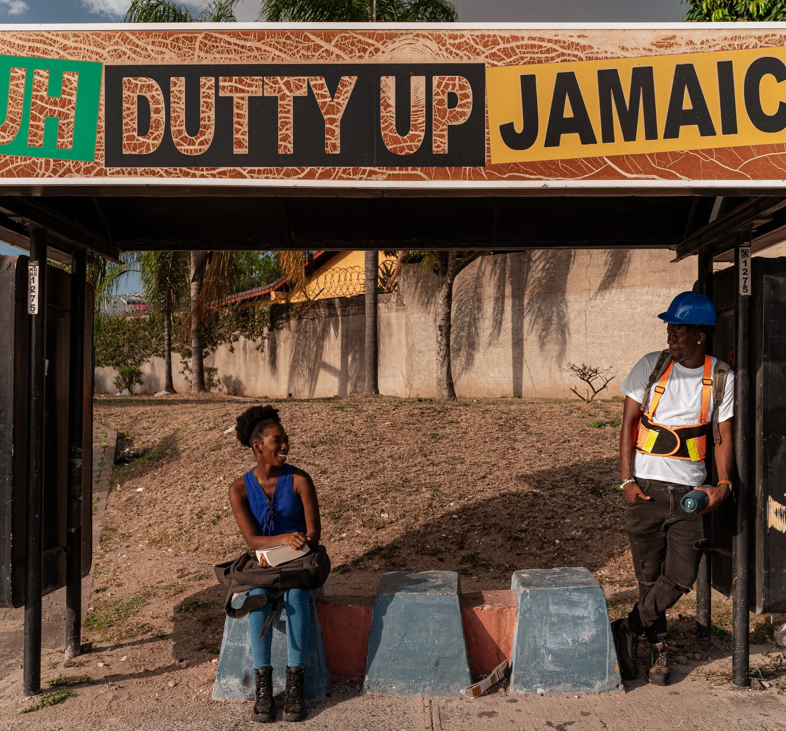 A young woman sits on a concrete barrier at a bus stop, smiling and holding a notebook, while a man in construction gear, wearing a blue hard hat and safety harness, leans against the shelter frame holding a water bottle. The bus stop sign reads 'DUTTY UP JAMAICA.'