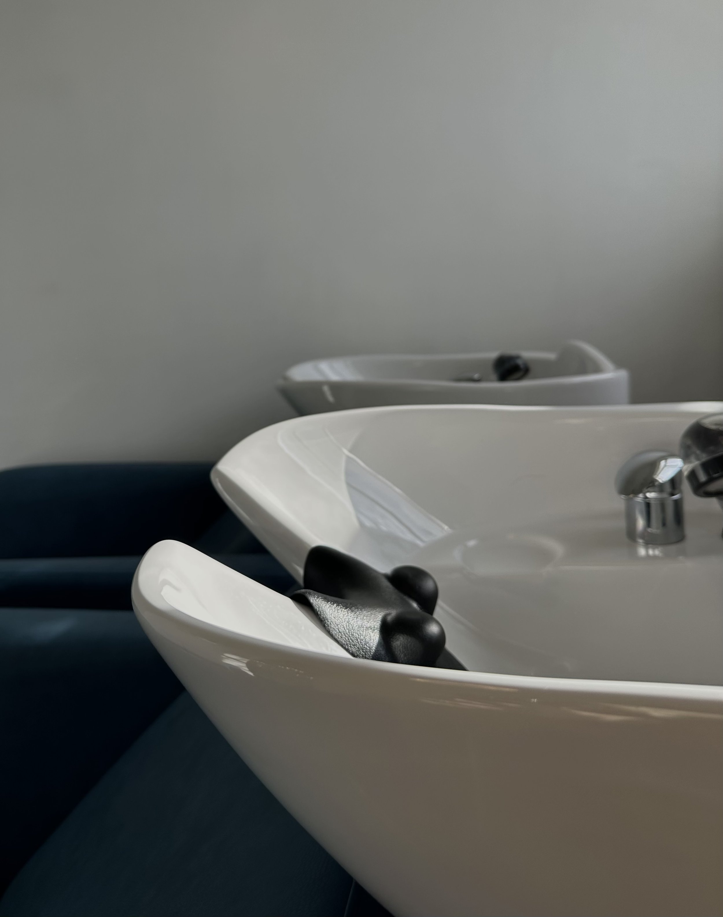 Two white salon washing sinks with black hair clips resting on the edge of the near sink, against a plain light wall background.