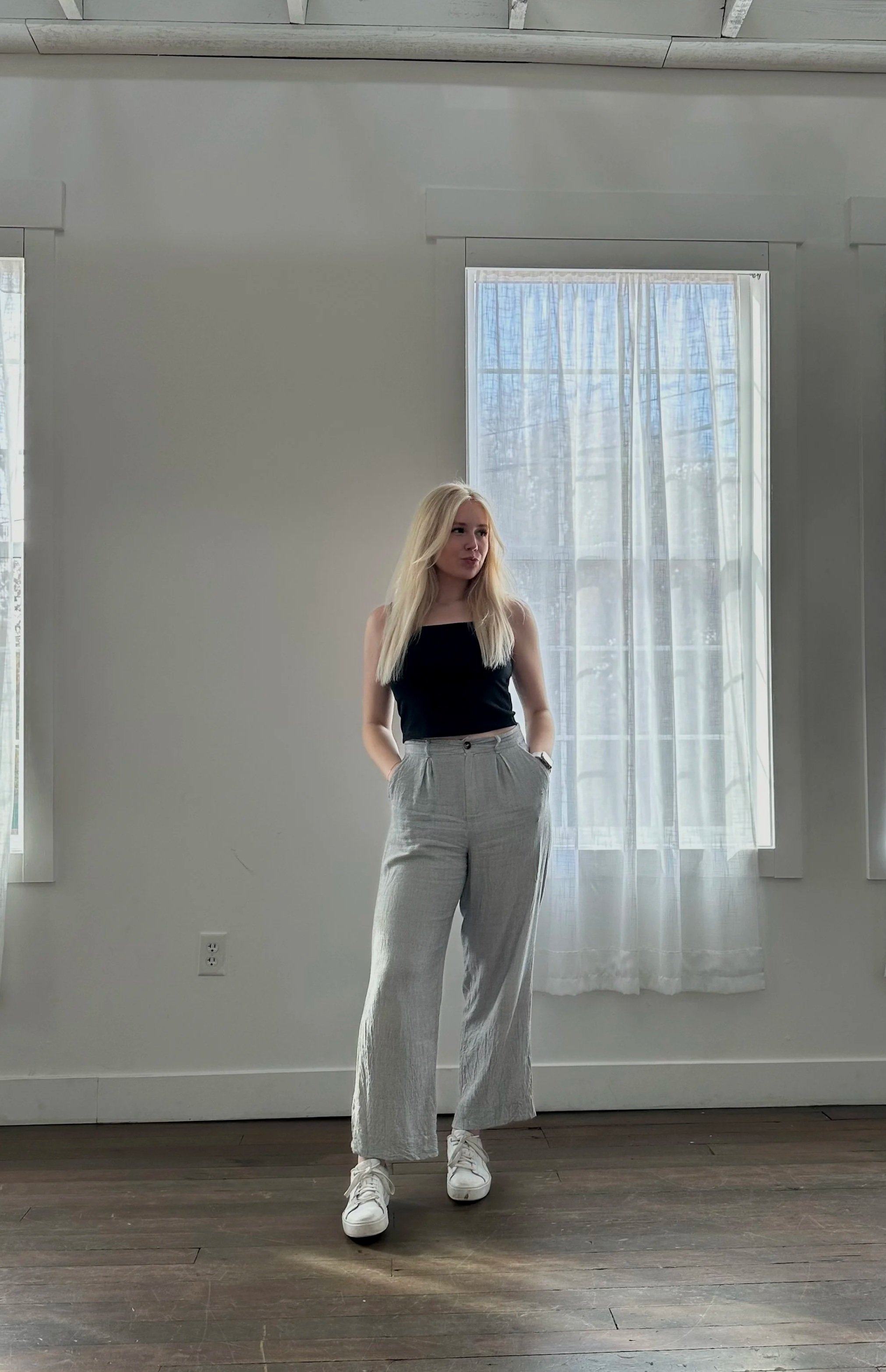 A young woman with long blonde hair stands in a room with white walls and dark hardwood floors, wearing a black sleeveless top, light-colored wide-leg pants, and white sneakers. She looks to the side with hands in her pockets, in front of a large window with white curtains that let in natural light.