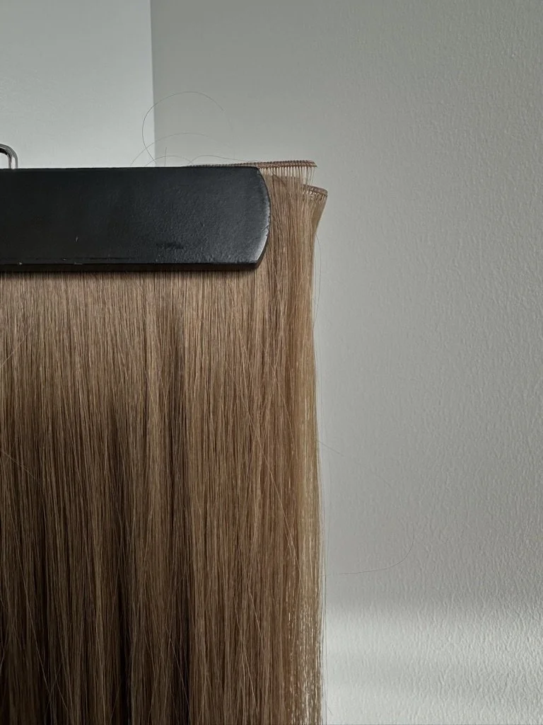 Close-up of hair extensions attached to a black clip on a wooden board, with a plain wall in the background.