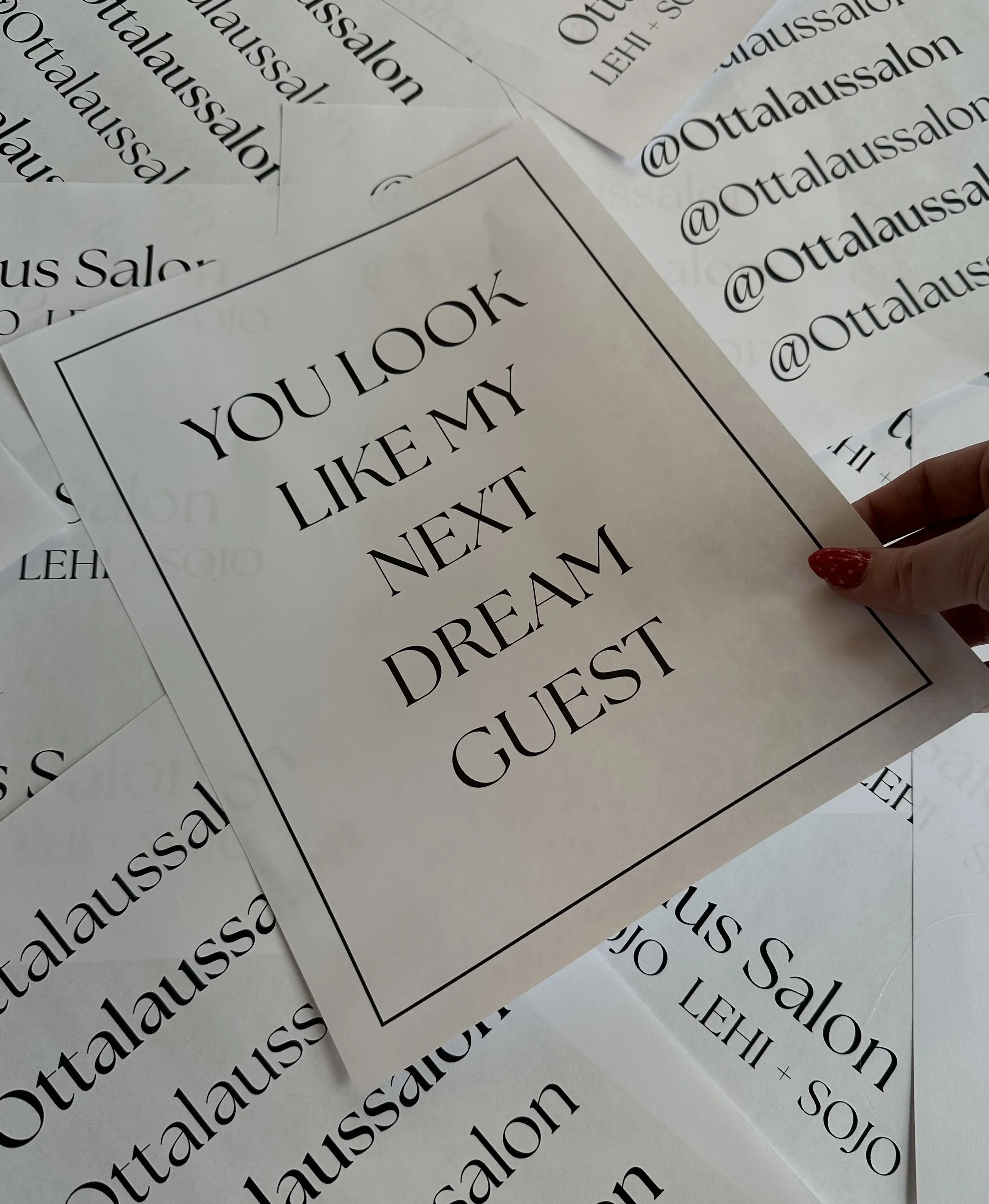 A person holding a sign that says, 'You look like my next dream guest,' with multiple printed papers on a surface, promoting a salon or beauty business.