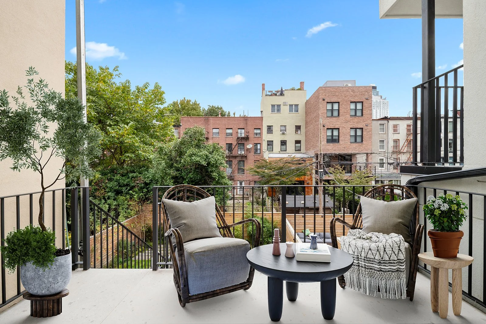 A balcony with two armchairs, a small round table with decorative vases and books, and potted plants, overlooking a view of city buildings and trees under a blue sky.