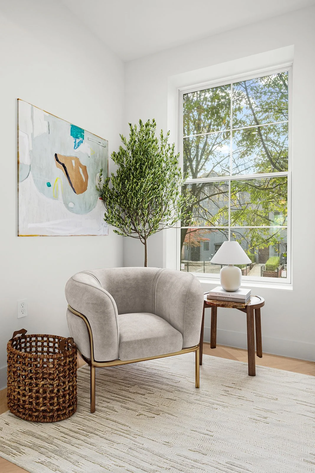 Bright living room corner with a large window, a modern beige armchair with gold legs, a small wooden side table with a white lamp and books, a potted tree, wall art, and a woven basket on a light-colored rug.