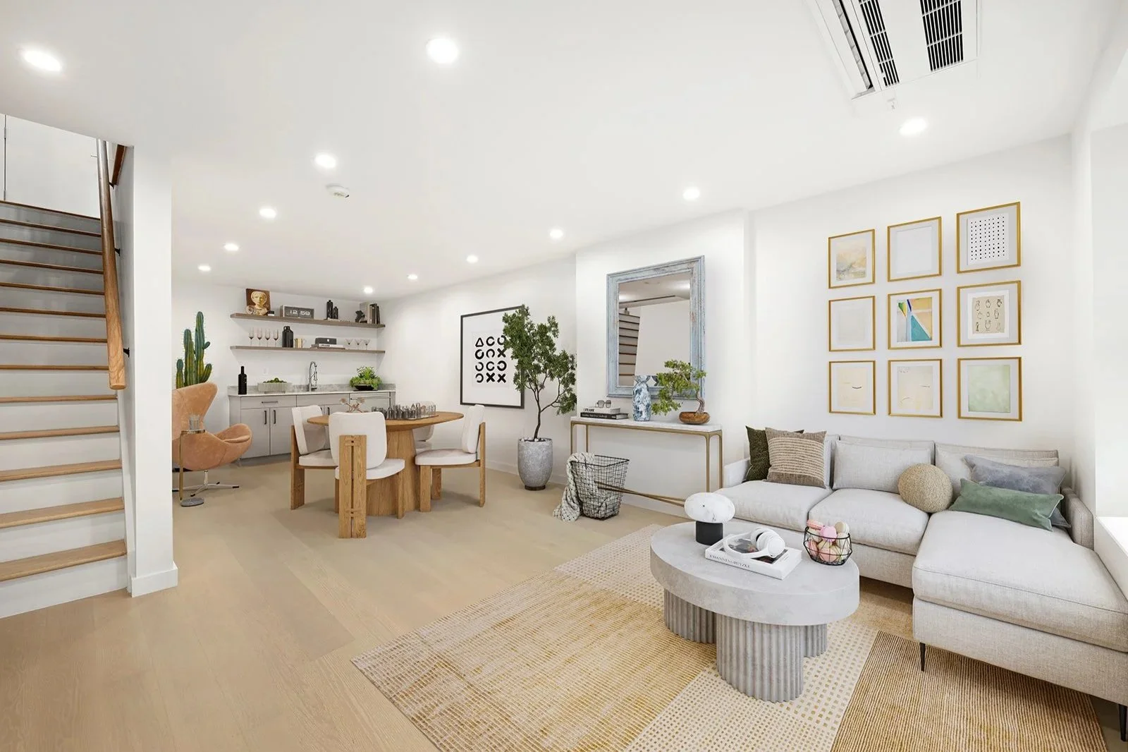 Bright, modern living room and dining area with white walls, wooden floors, a white sectional sofa with assorted pillows, a round marble coffee table, and a wall with framed artwork. The dining area has a round wooden table with white chairs, a wall-mounted shelf with decorative items, and a side cabinet beneath. A staircase with wooden steps is visible on the left.