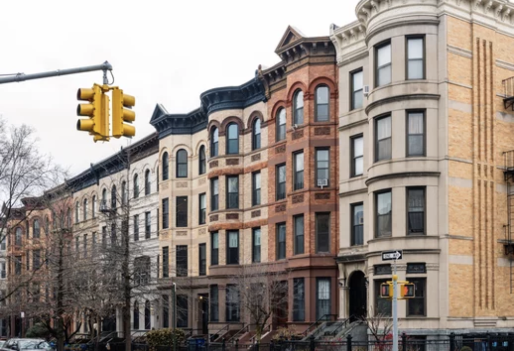 Row of historic brownstone and brick townhouses on a city street with traffic lights.